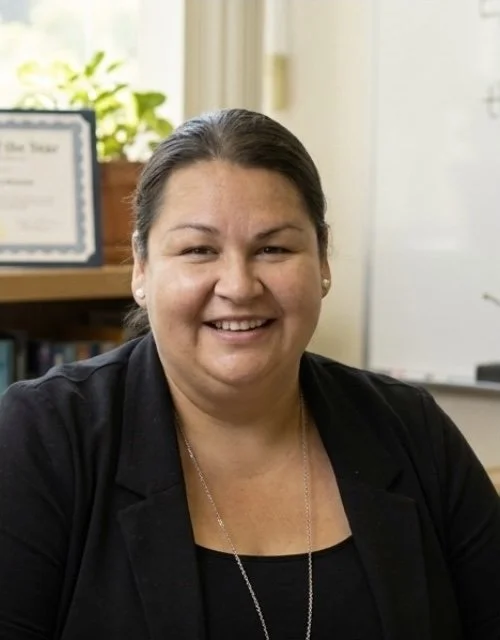 Smiling woman with dark hair in a bun, wearing a black blazer, sitting at a desk in an office with a diploma and green plant in the background.