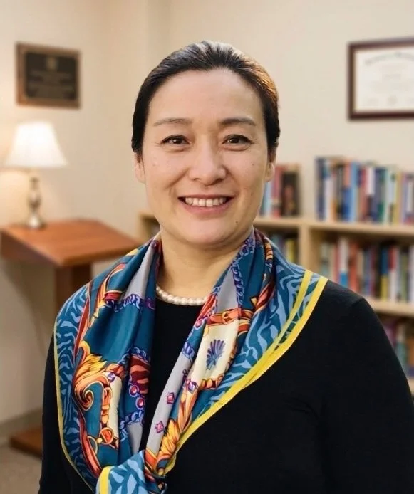 A smiling woman with dark hair pulled back, wearing a colorful silk scarf and pearl necklace, standing in an office or library with bookshelves and a lamp in the background.