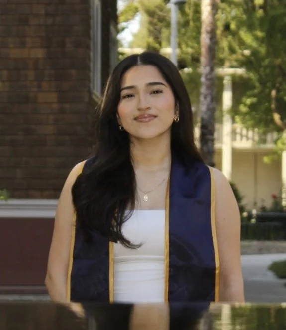 Young woman with long dark hair wearing a white top and a blue graduation stole with gold trim, standing outdoors in front of a brick building and trees.