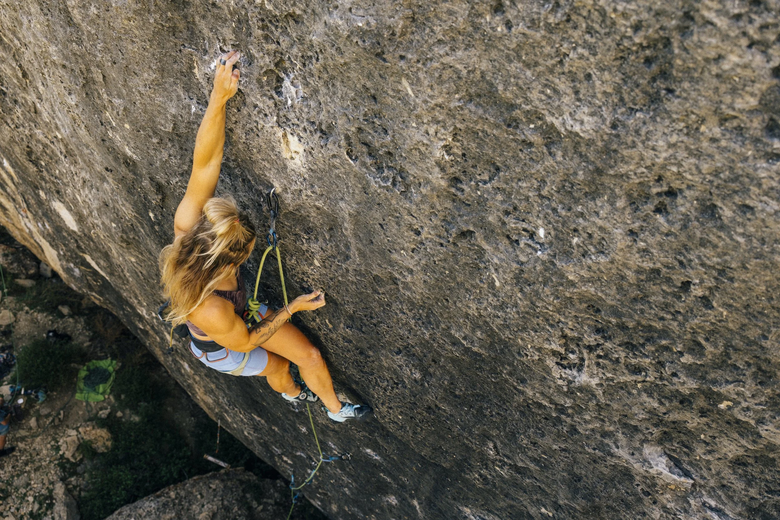 A woman wearing a face mask, gray tank top, and jeans climbing an indoor bouldering gym wall with colorful holds.