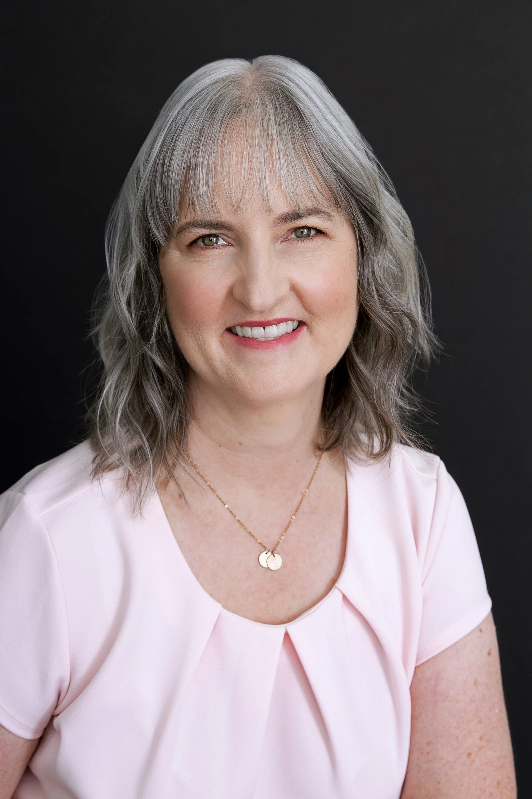 Smiling woman with brown hair and bangs wearing red lipstick and a pink top against a blurred outdoor background.