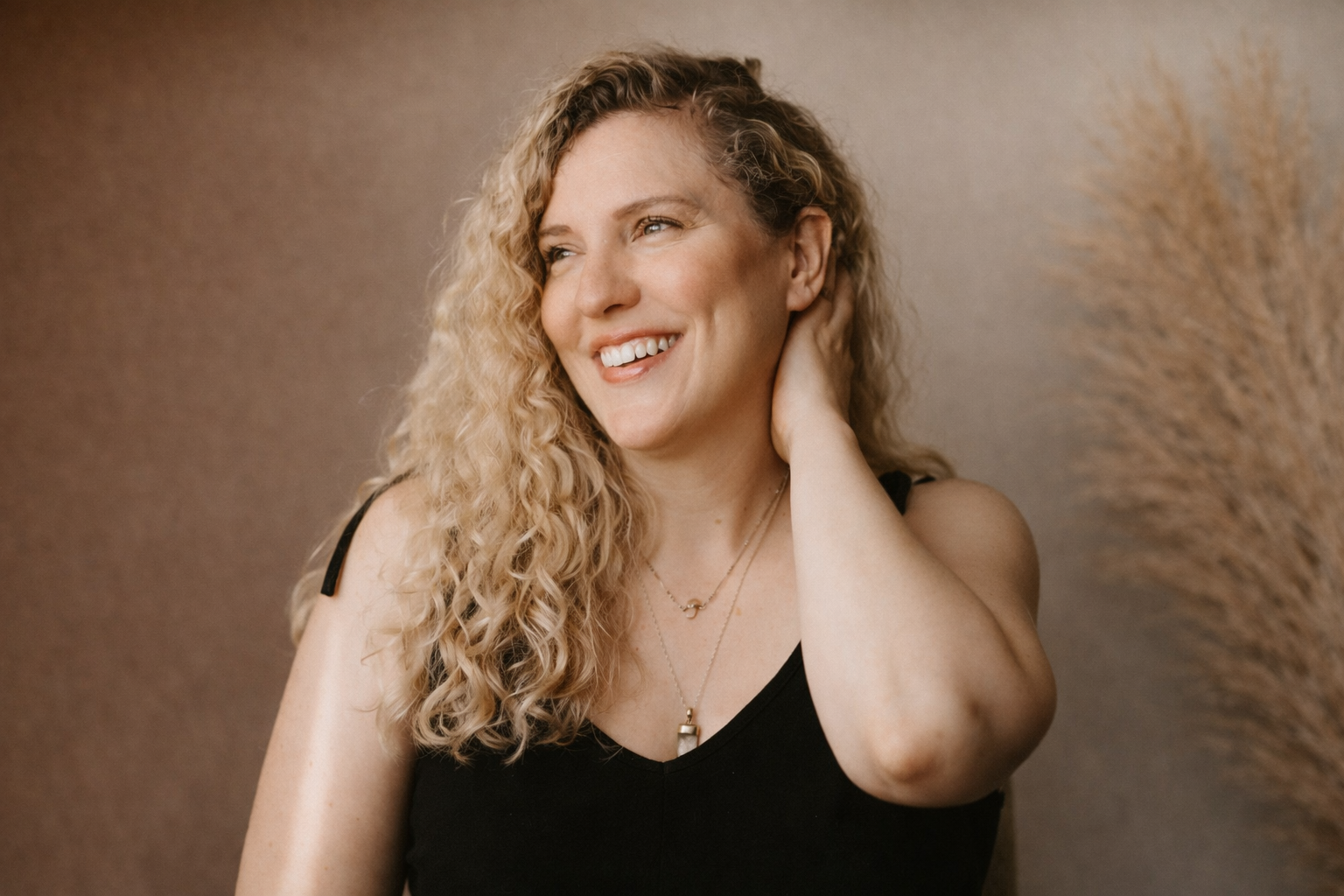 A woman with curly blonde hair, wearing a black top, smiling and touching her neck, with a beige background and decorative dried grass on the right.