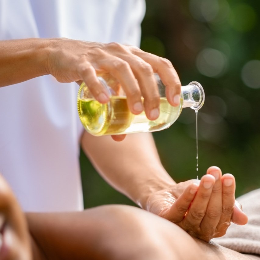 Two hands holding a clear bottle with a yellow massage oil in it.  Oil is being poured out of the bottle into one hand that is resting on a woman's back who is about to get a massage.