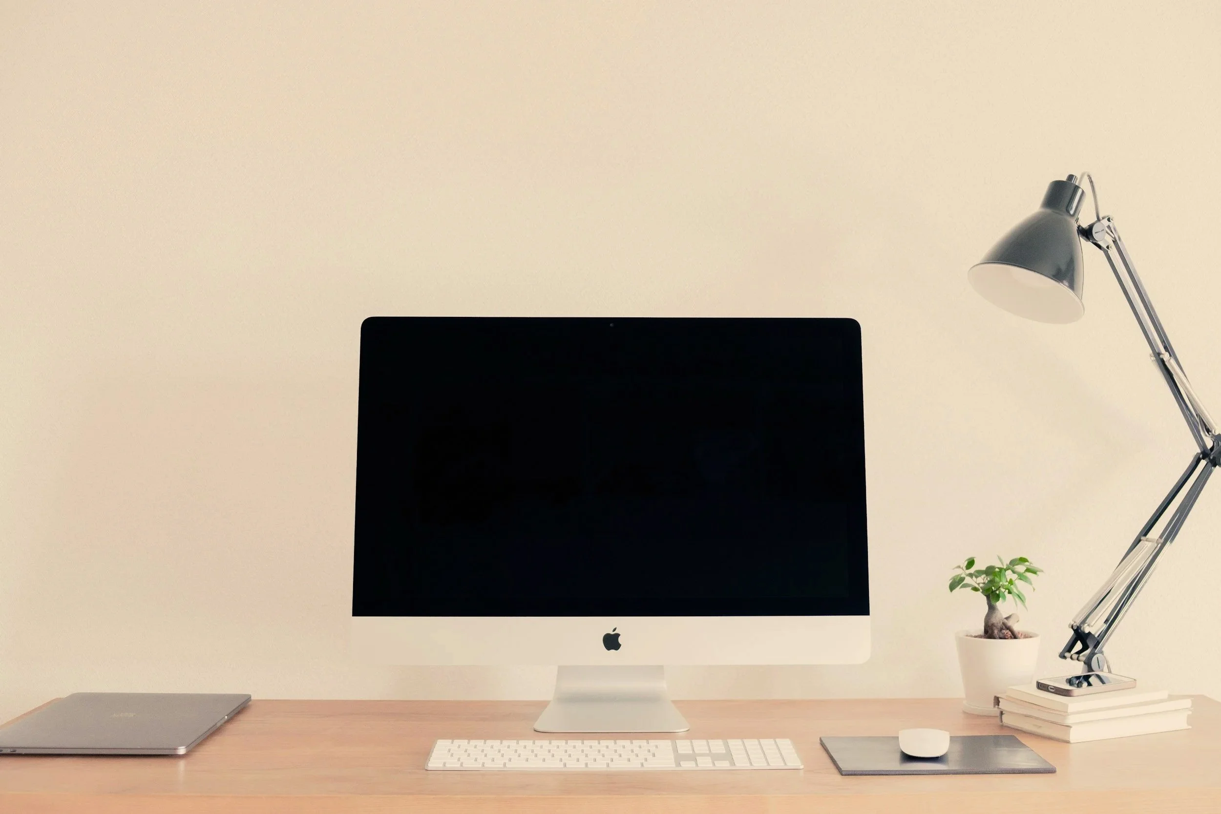 Desk setup with an Apple iMac computer, a closed laptop, a wireless keyboard, a mouse on a desk mat, a potted plant, a stack of books, and a desk lamp.