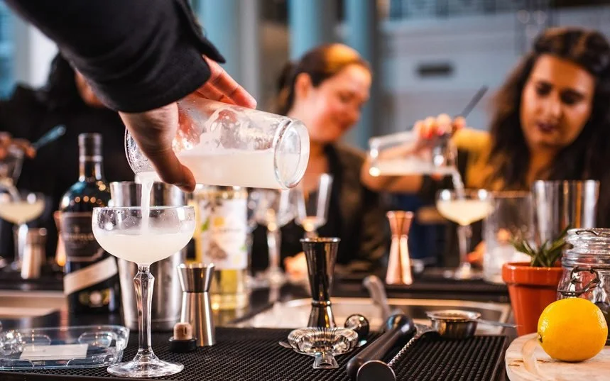 A bartender pouring a white cocktail into a glass at a bar with people in the background.