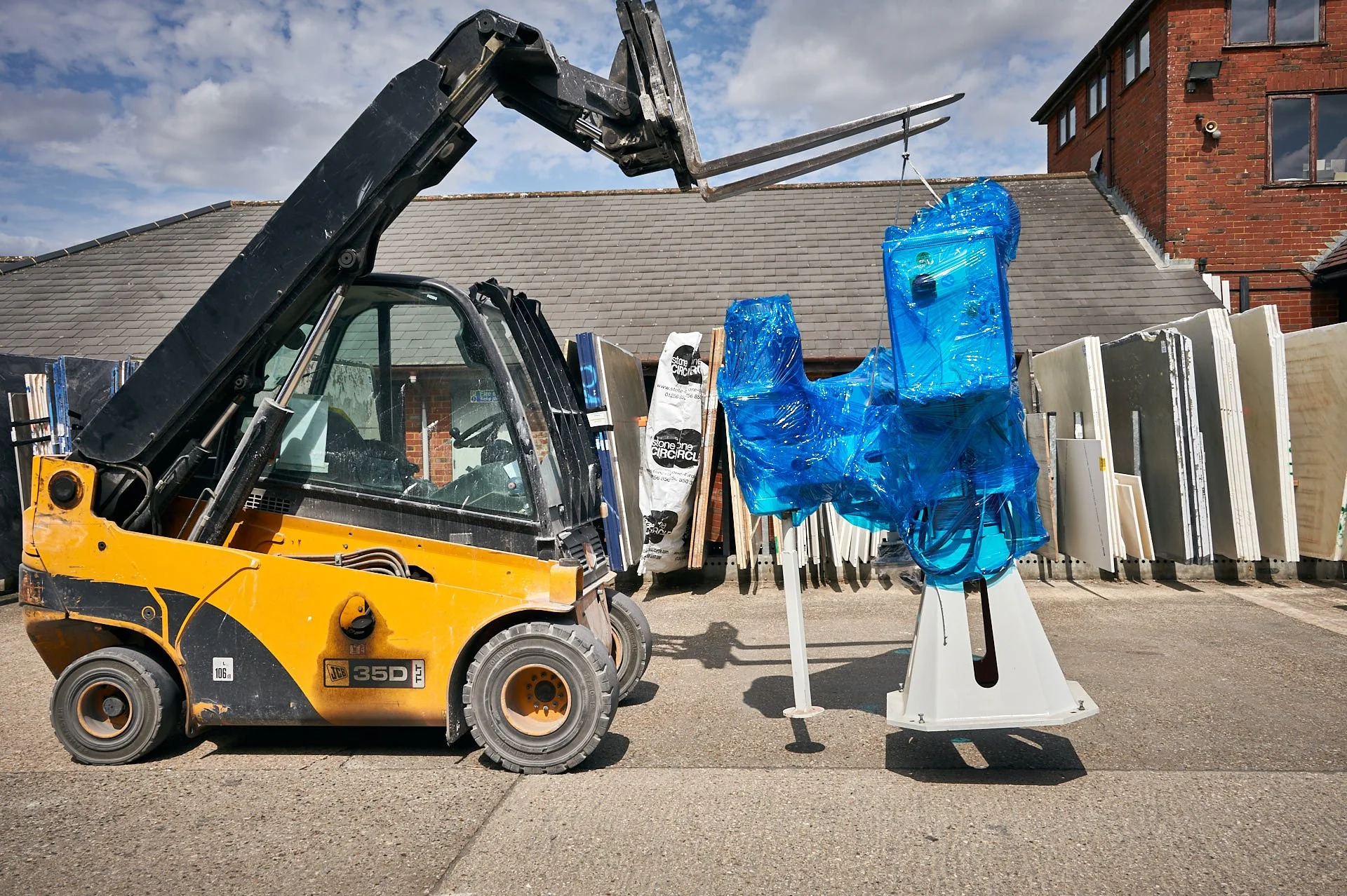 A yellow telehandler lifts the replacement polishing machine, wrapped in blue film, beside rows of upright stone slabs.