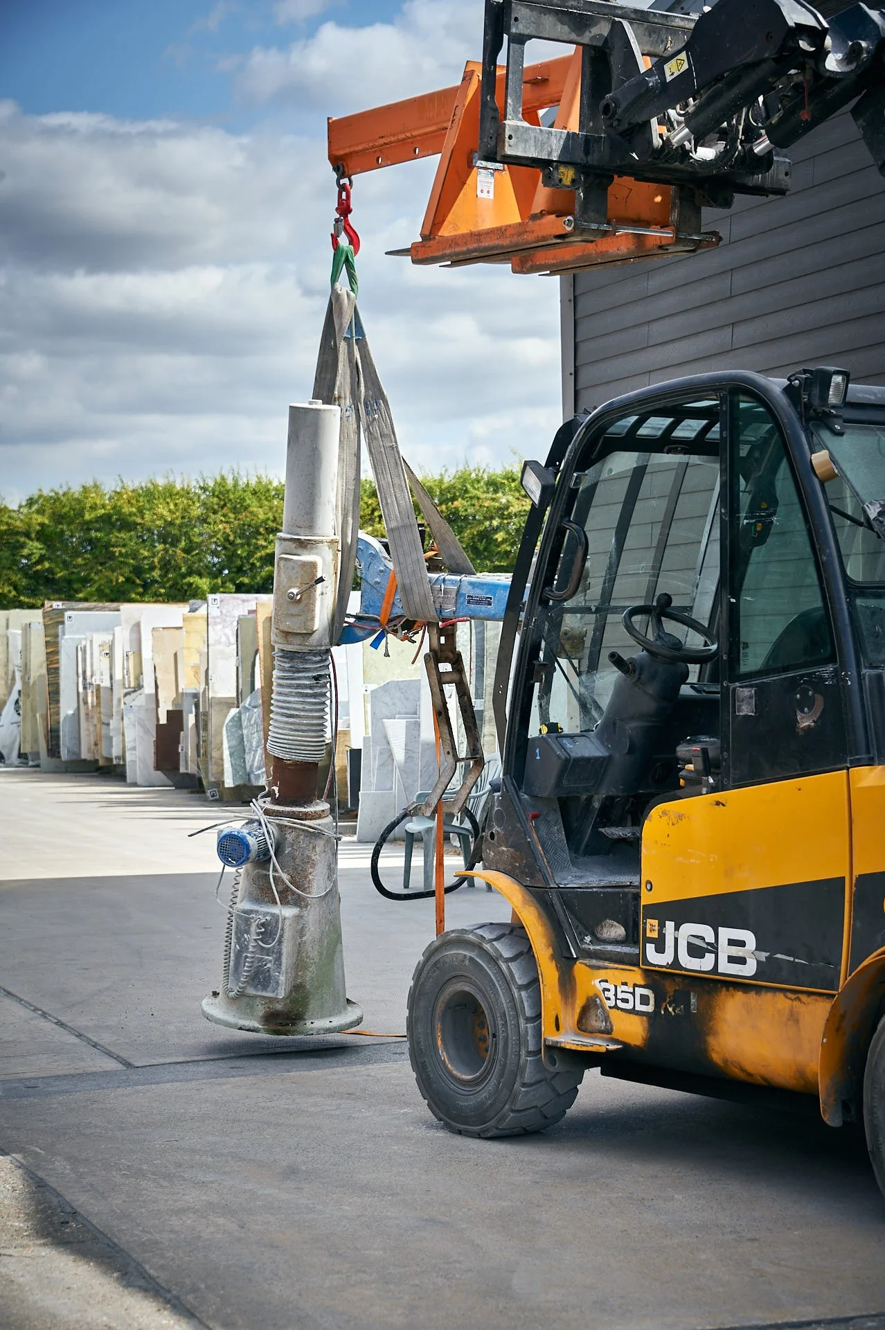 The old Jenny Lind polishing machine hangs from lifting straps beside a yellow telehandler in the stone yard.