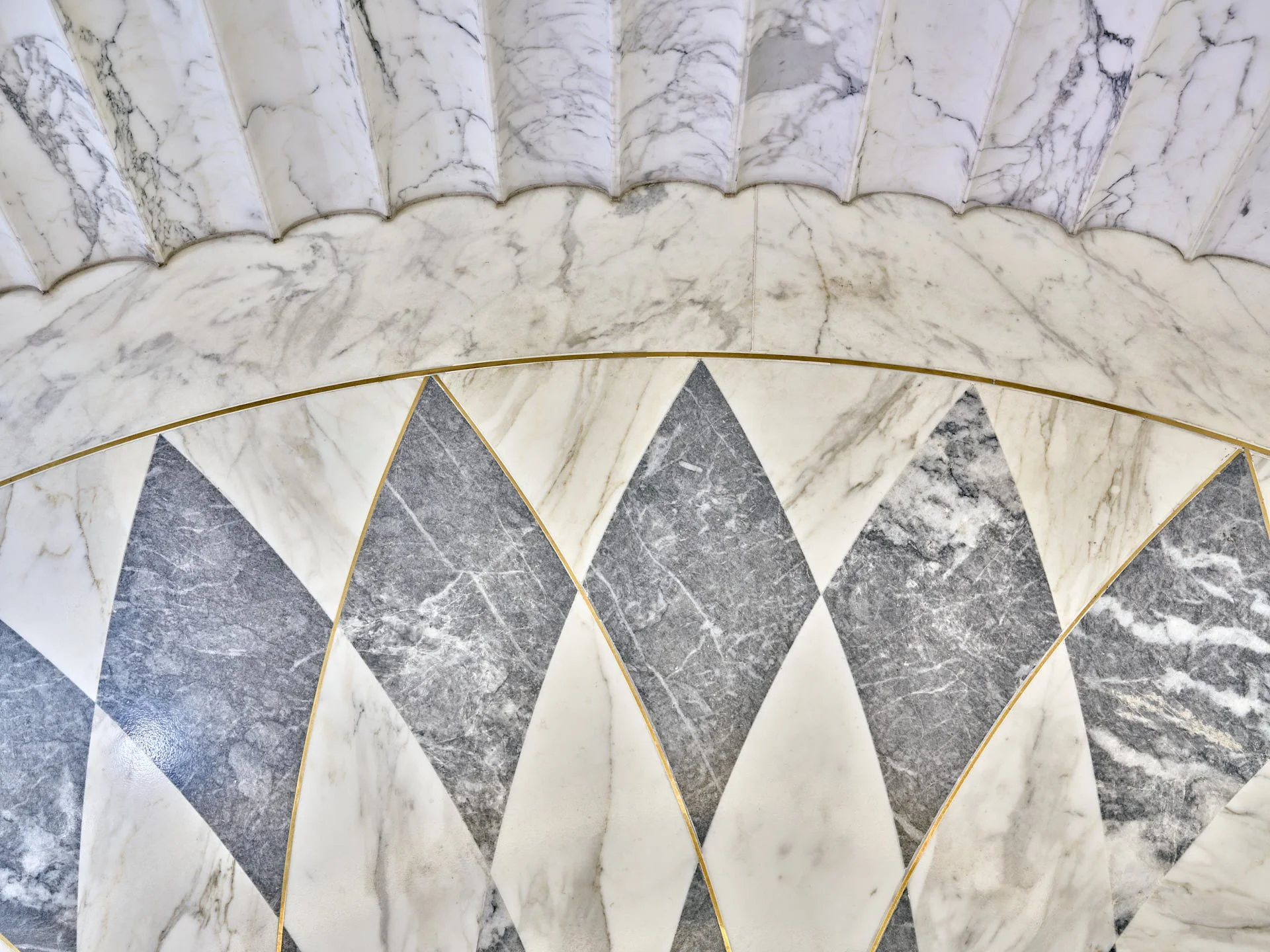 Close view of alternating pale and blue-grey stone pieces meeting fluted marble wall panels at a curved brass border. Fabrication and installation by stoneCIRCLE - photo Marco Joe Fazio Studio.