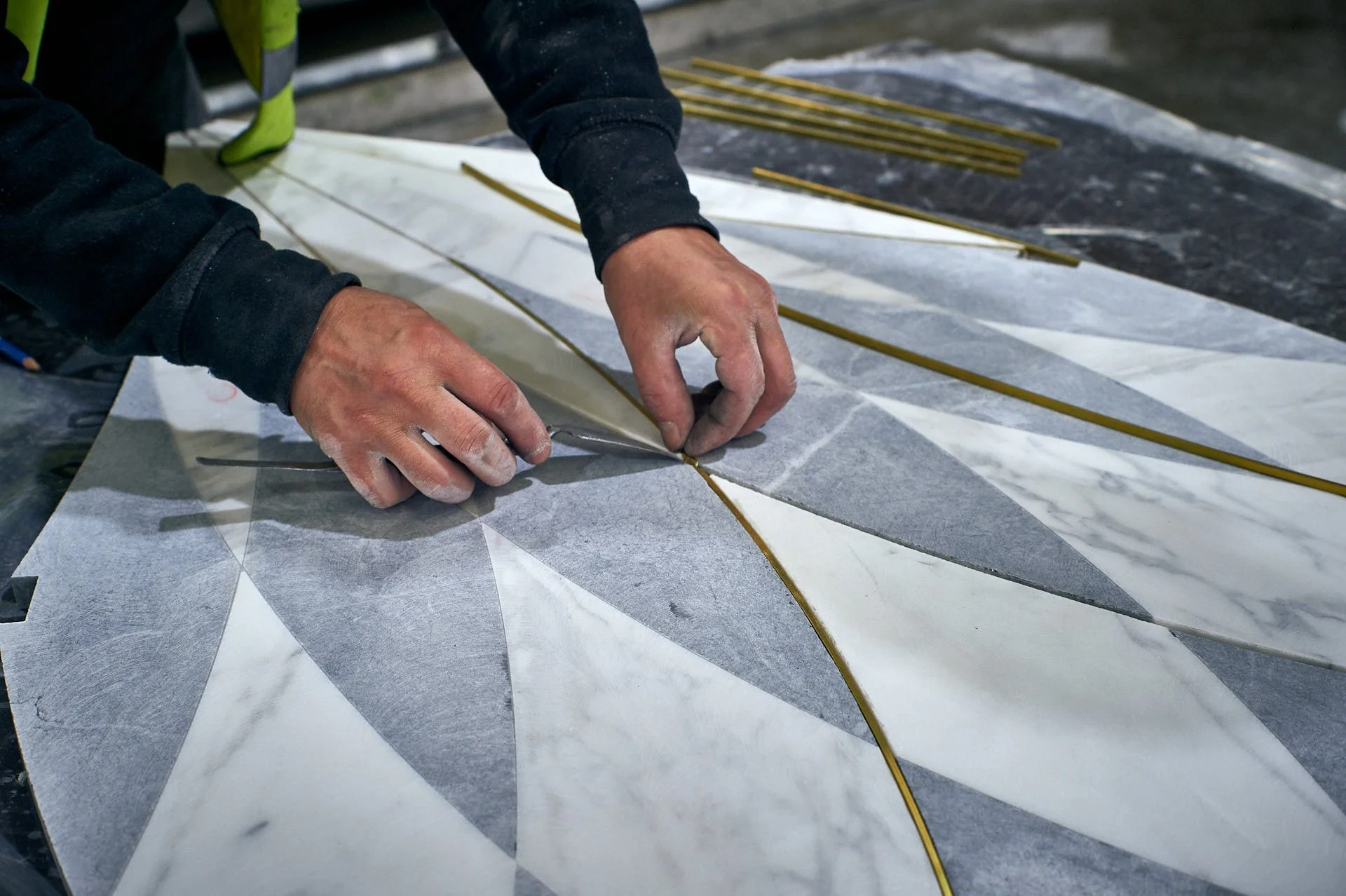 Close view of a craftsperson fitting a narrow brass strip by hand into a curved groove between pale and blue-grey stone pieces. Fabrication by stoneCIRCLE - photo Marco Joe Fazio Studio.