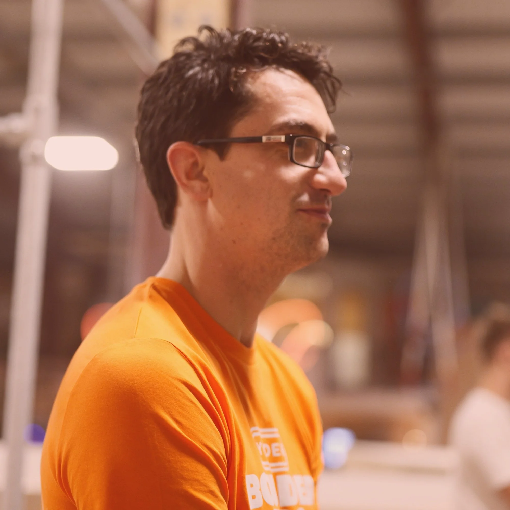 Side profile of a man with dark hair, glasses, and wearing an orange shirt in an indoor setting with warm lighting.
