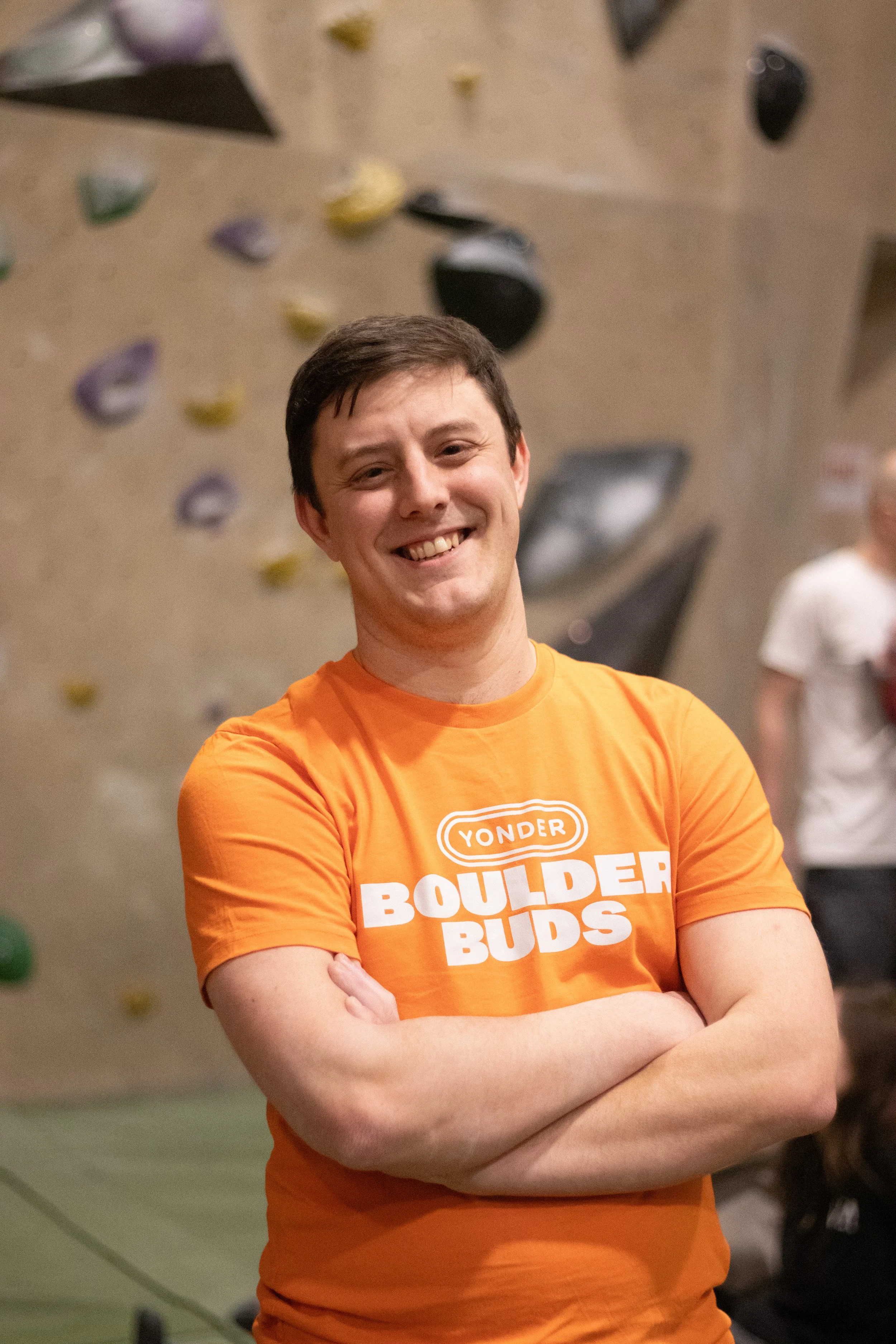 A smiling man with short dark hair, wearing an orange t-shirt that says "Yonder Boulder Buds," standing with his arms crossed inside a climbing gym, with a climbing wall and some blurred individuals in the background.