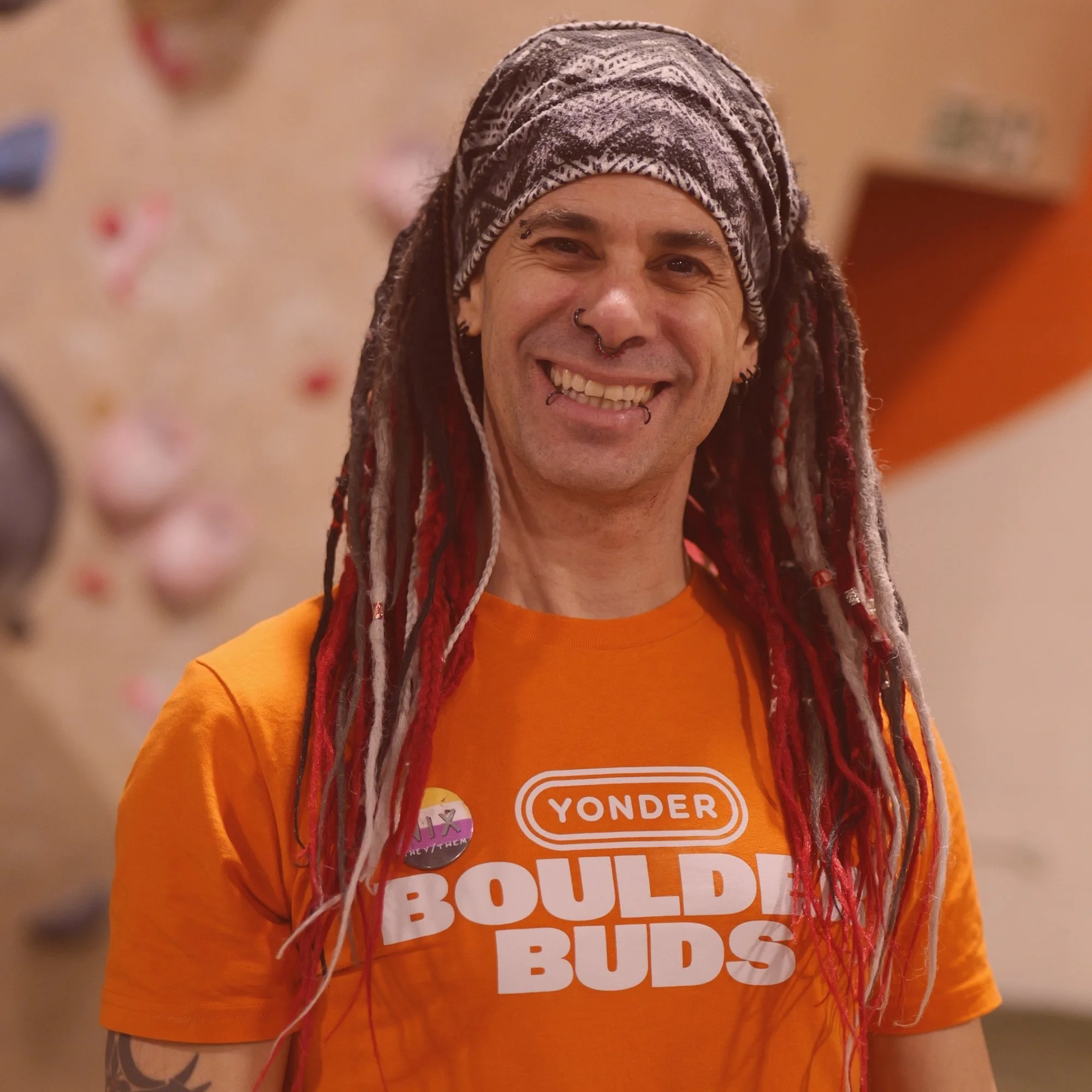 A smiling man with long dreadlocks and various facial piercings wearing an orange shirt with the text 'Yonder Boulder Buds' and a small button on his chest, standing in front of a blurred background with climbing holds.