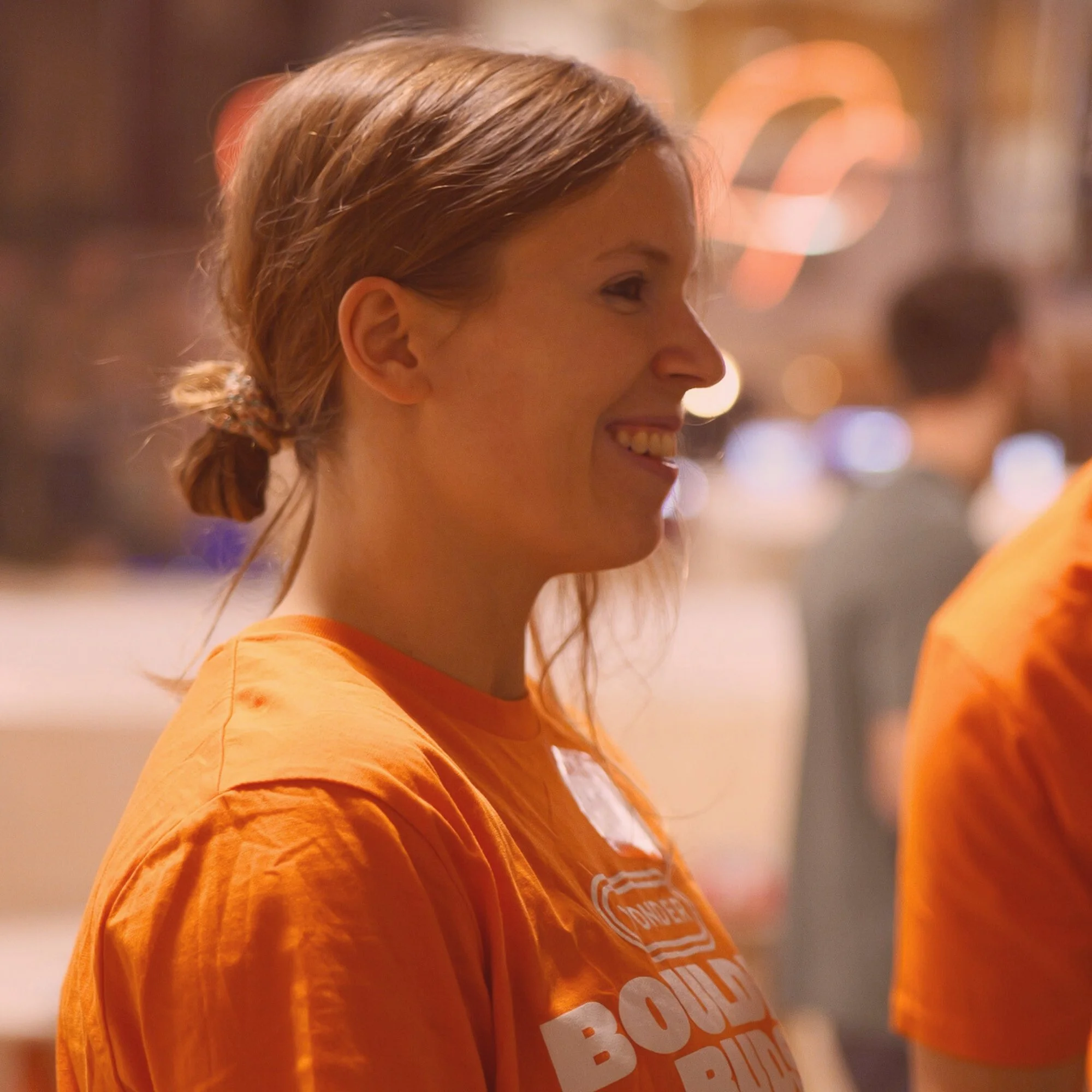 A young woman with light brown hair tied back in a bun, smiling, wearing an orange T-shirt with white text, in a warmly lit indoor setting with blurred background figures.