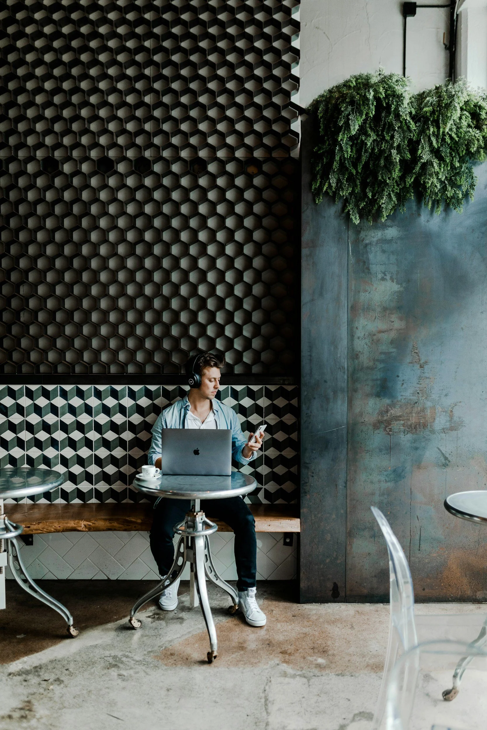 Person som sitter ved et kafébord med en bærbar datamaskin og mobiltelefon i en moderne, stilfullt innredet kafé med heksagonalt mønstrede vegger og hengende planter.