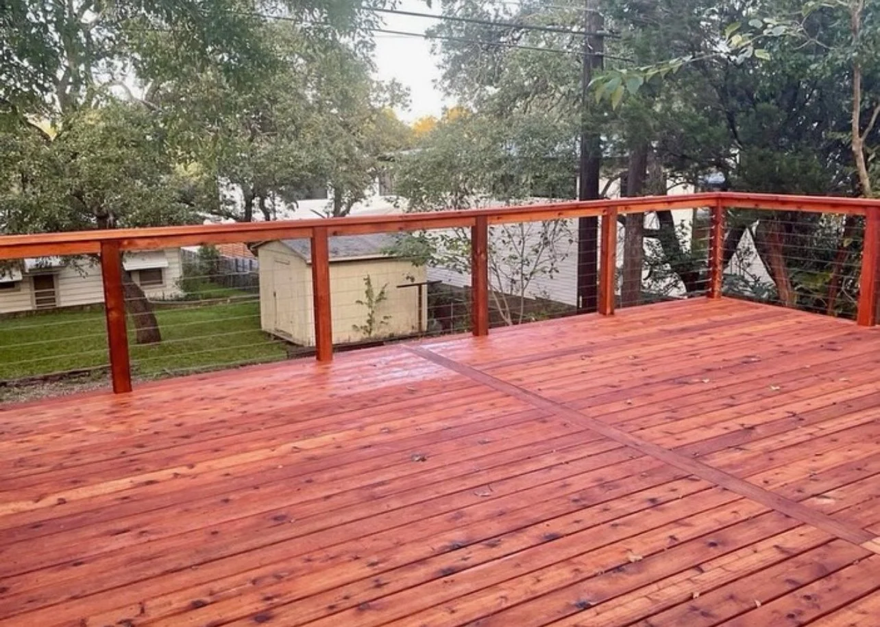 Red-stained wood deck with railing in a tree-filled yard.