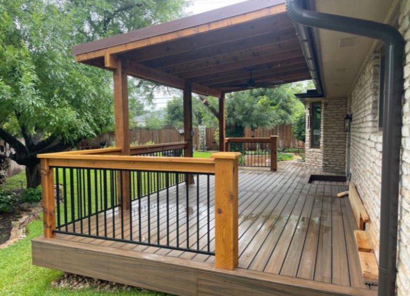 Covered deck with wood posts and black metal railing beside a home.