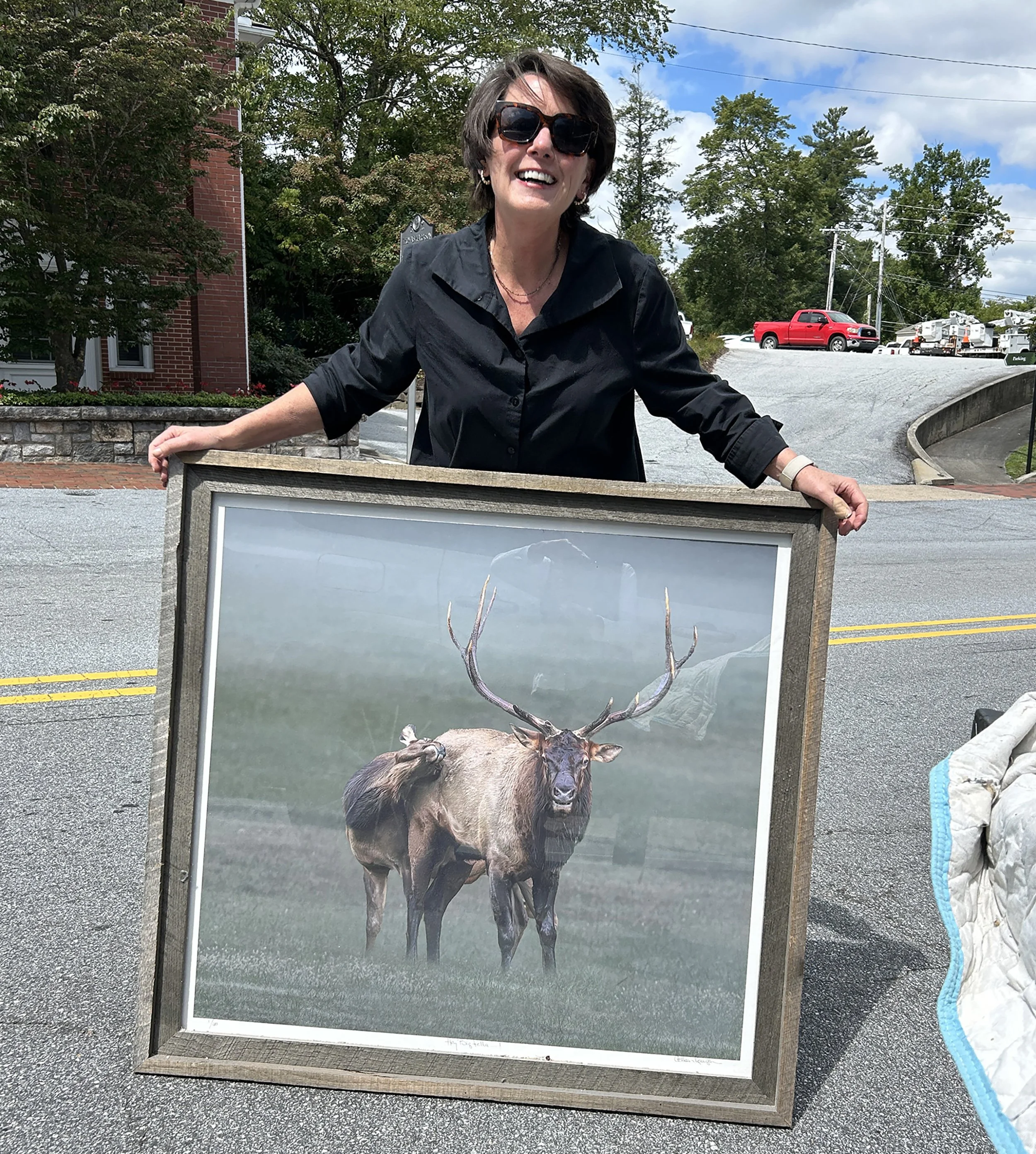 A woman with short dark hair, wearing sunglasses and a black blouse, smiling and holding a large framed photo of a moose with impressive antlers, outdoors on a street.