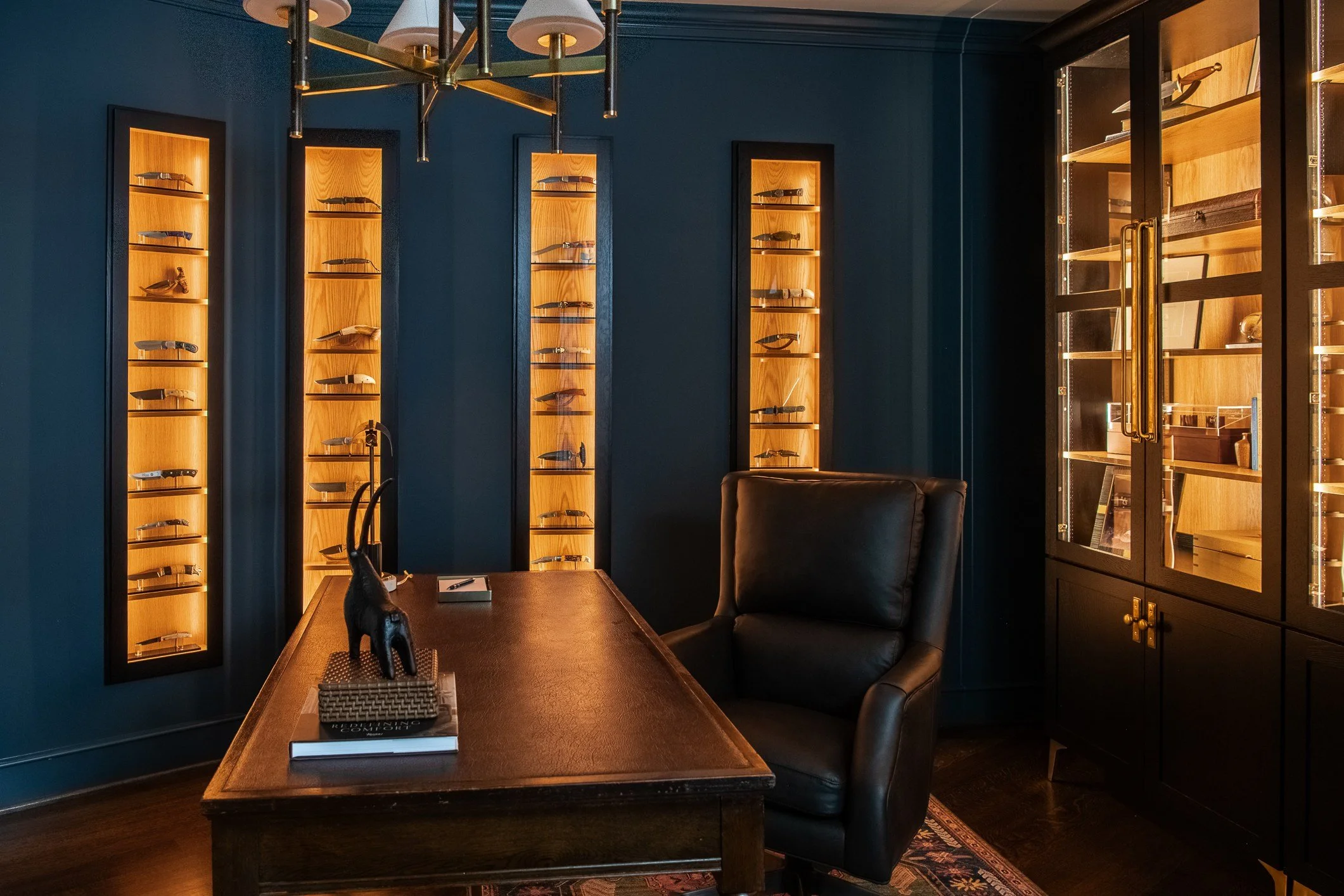 A room with dark blue walls, wooden display cases with lighting showcasing knives, a wooden desk with a black leather chair, and a decorative sculpture of a black animal on the desk.