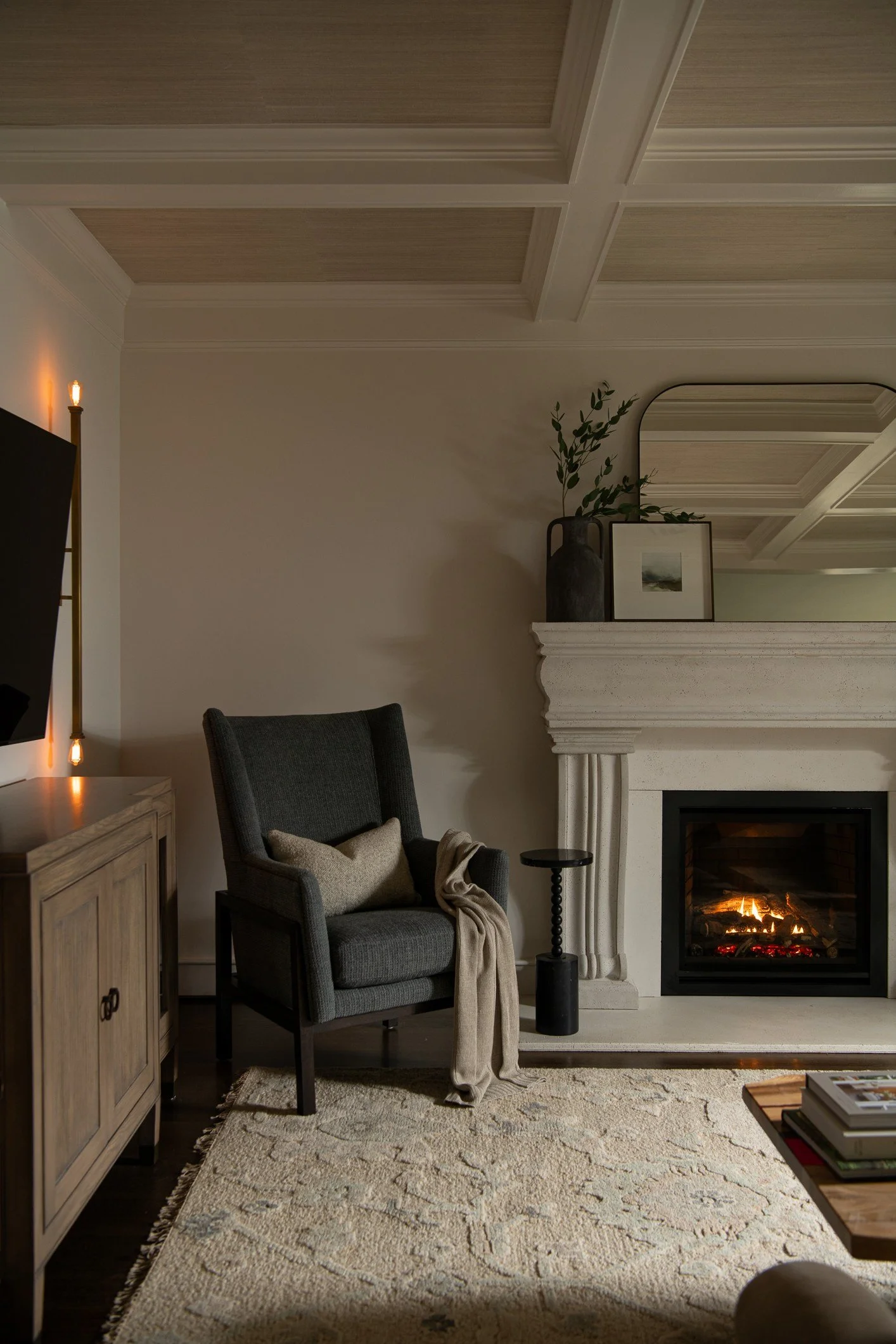 A cozy living room corner featuring a black high-back armchair with a beige cushion and throw, a beige patterned area rug, a white fireplace with decorative trim and a fire burning, and a wooden sideboard. A black vase with greenery and a framed pict