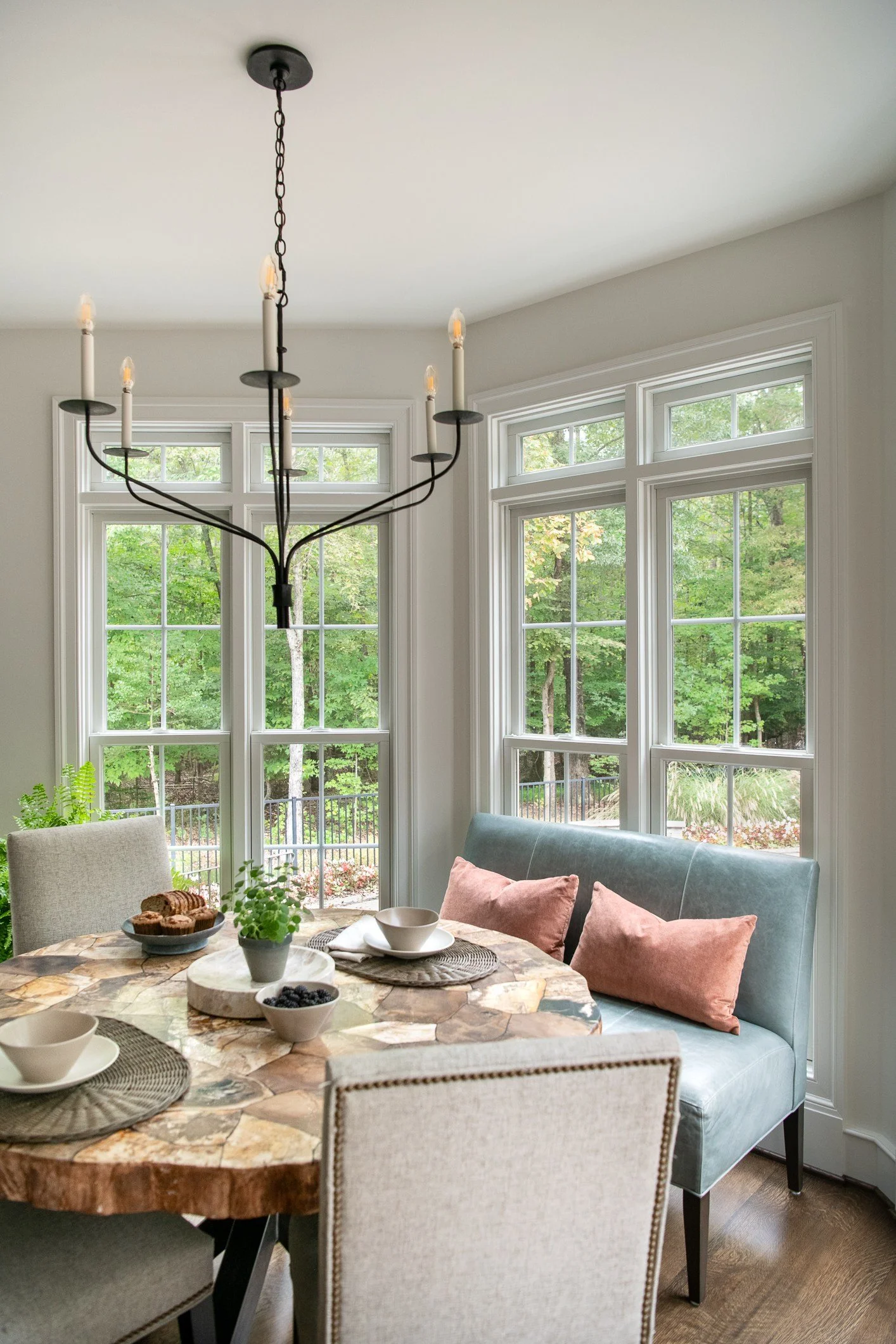 A dining room with a round stone table, beige and teal chairs, a light fixture with candles, and large windows showing a green outdoor landscape.