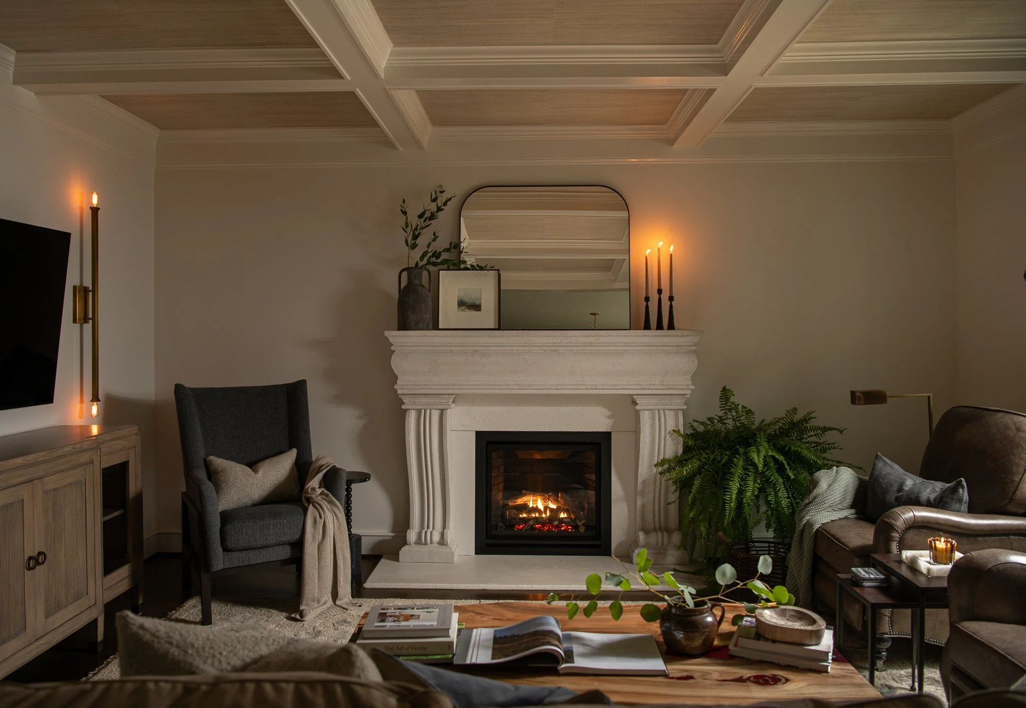 Cozy living room with a fireplace, armchair, leather sofa, plants, books, and warm lighting