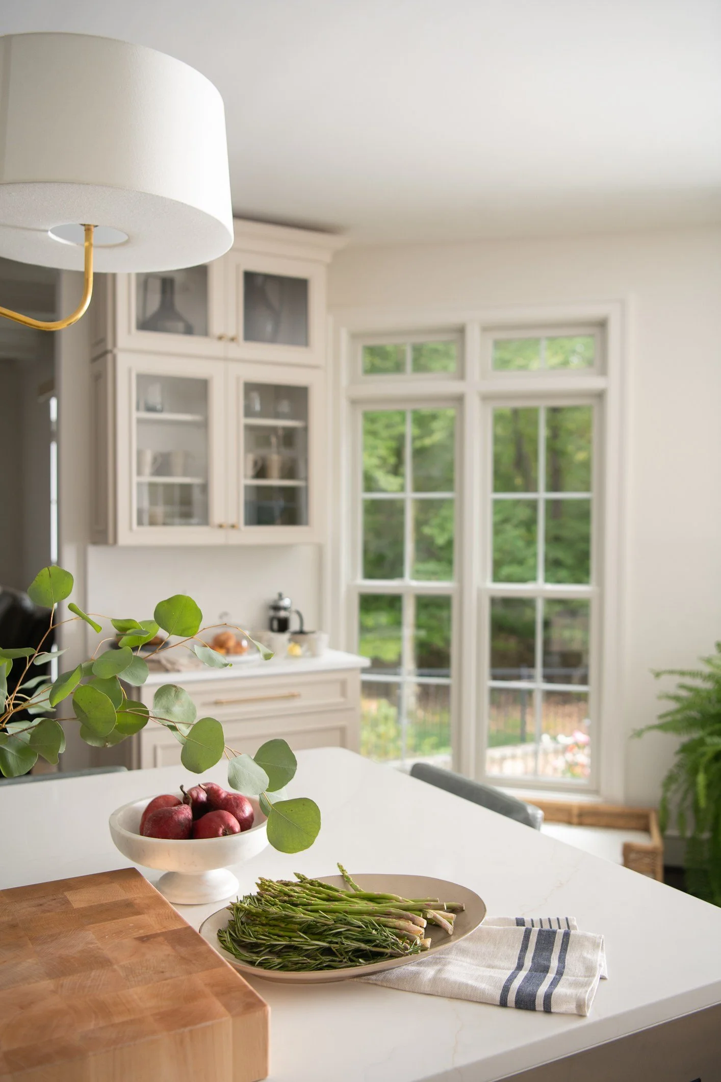 A bright kitchen with large windows, white cabinetry, a white countertop, a bowl of red apples, and a plate of fresh green asparagus, with a wooden cutting board and a striped cloth on the counter.