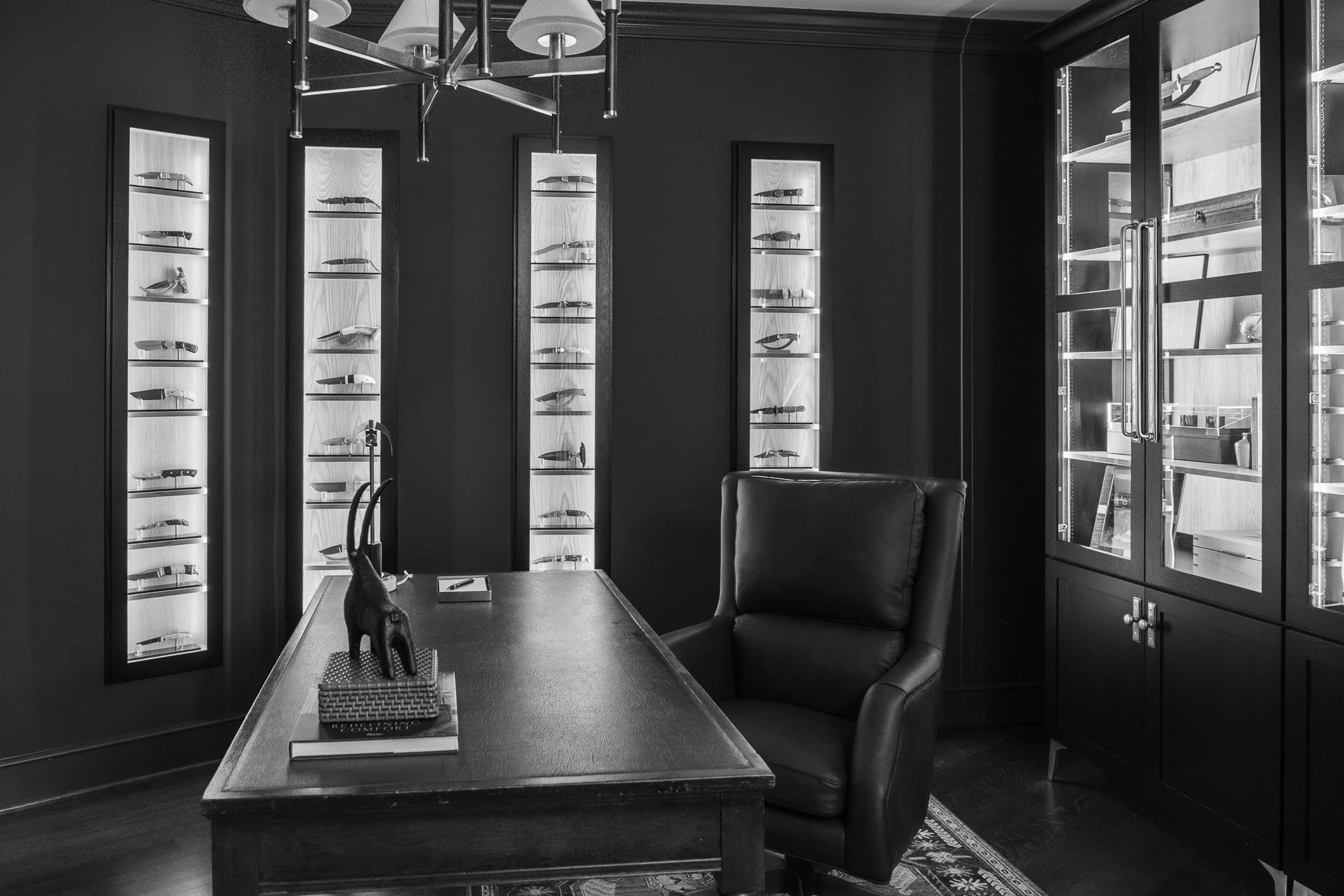 A black and white photo of a study or office with dark walls and wooden furniture. There is a large desk with a decorative animal figure and books on it, a tufted leather chair, tall display cabinets with glass doors holding knives or similar objects