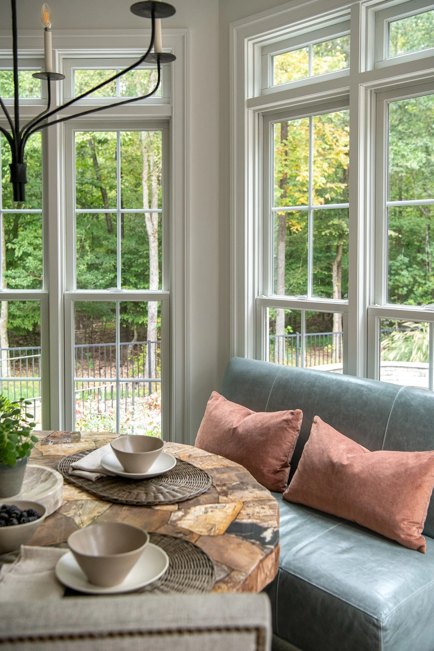 Bright dining nook with large sash windows showing green trees outside, a marble-top table set with bowls and plates, and a cushioned bench with pink pillows.