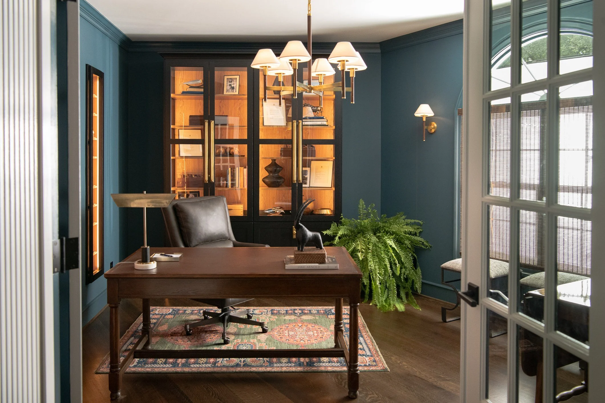 An office with a wooden desk, black leather chair, large green fern, and a cabinet with books and decorative objects. Light fixtures on the wall and chandelier above.