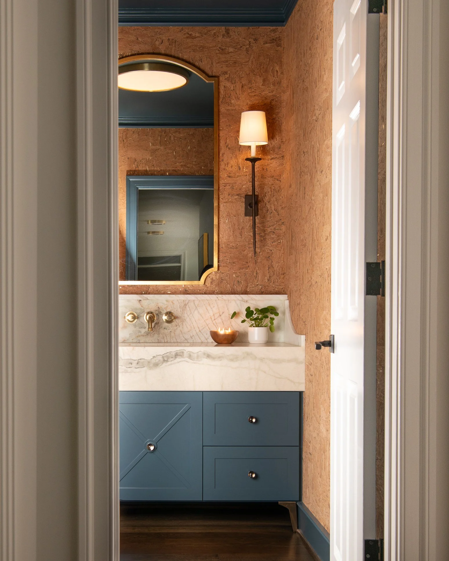 A bathroom with a small blue vanity, tan marble countertop, and a mirror. There is a wall-mounted lamp and a small potted plant with a bowl of candles on the countertop.