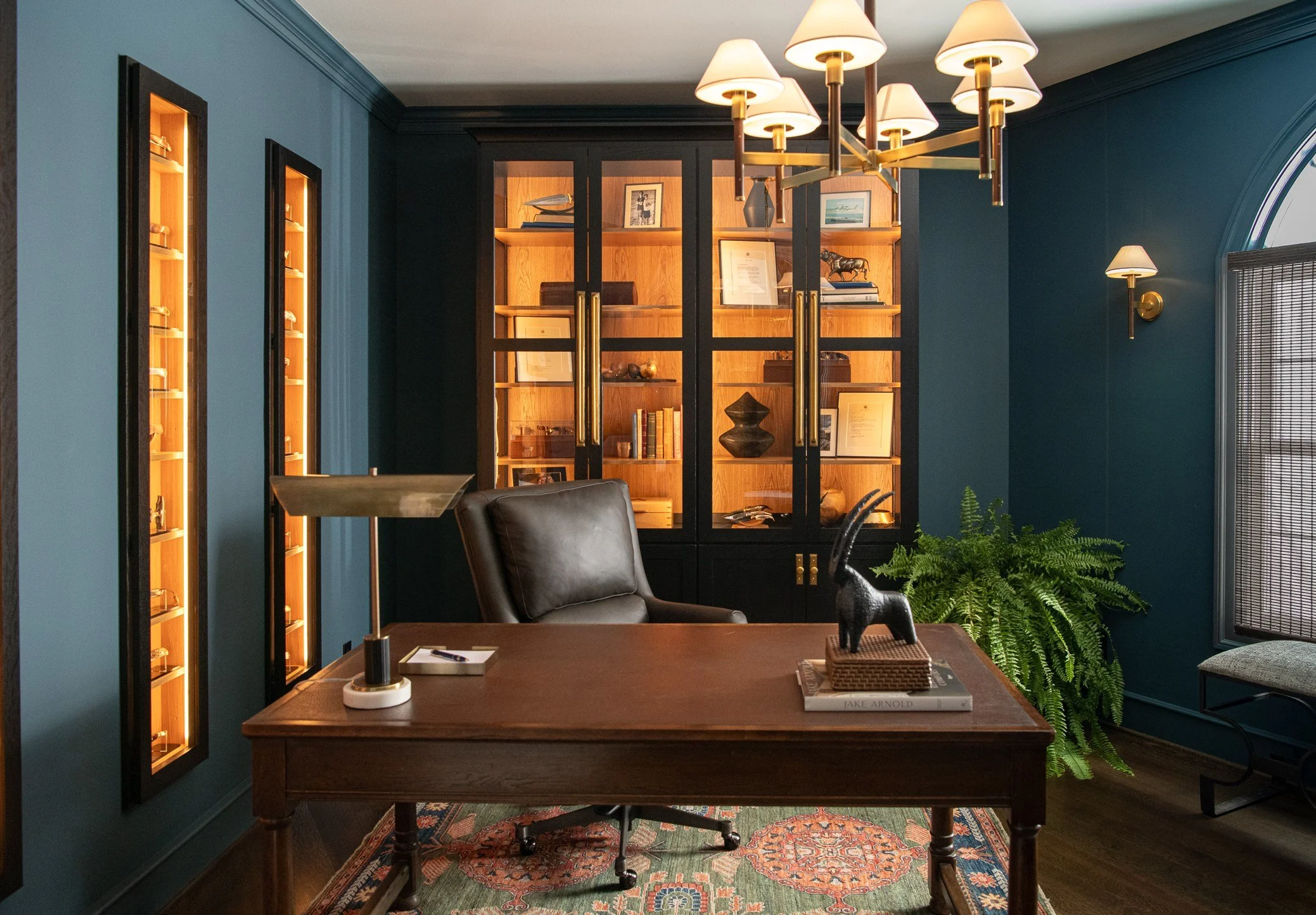 An office with a wooden desk, black leather chair, blue walls, and a large wooden cabinet with glass doors. The cabinet contains books and decorations. There's a fern plant in the corner, a window with blinds, a modern chandelier above the desk, and 