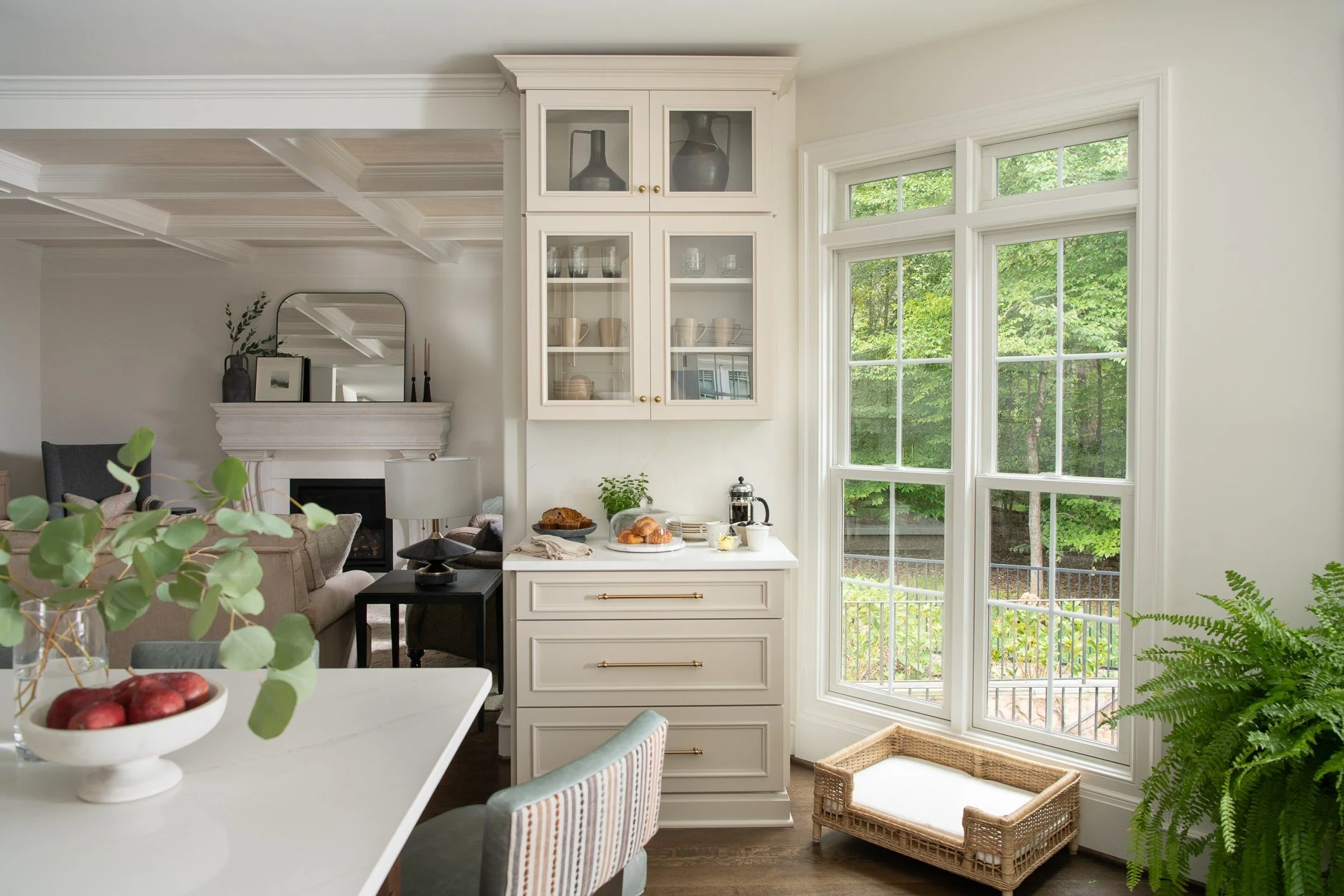 Bright kitchen corner with white cabinetry, large windows showing green trees outside, a wicker pet bed, and various decor items.