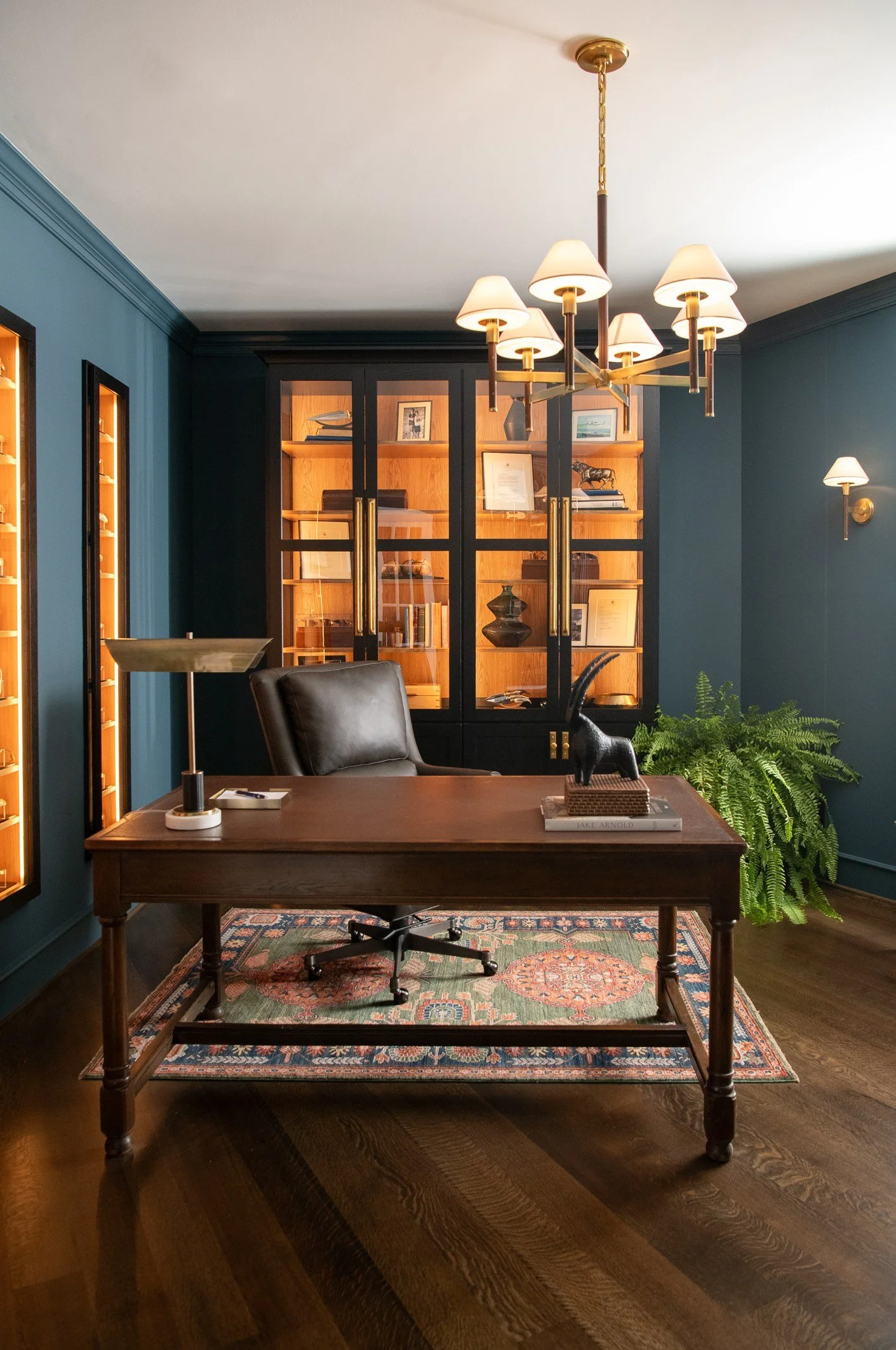 Modern home office with dark wood desk, leather chair, green fern plant, blue walls, wall-mounted lamps, and a glass-fronted bookshelf with framed pictures and decorative objects.