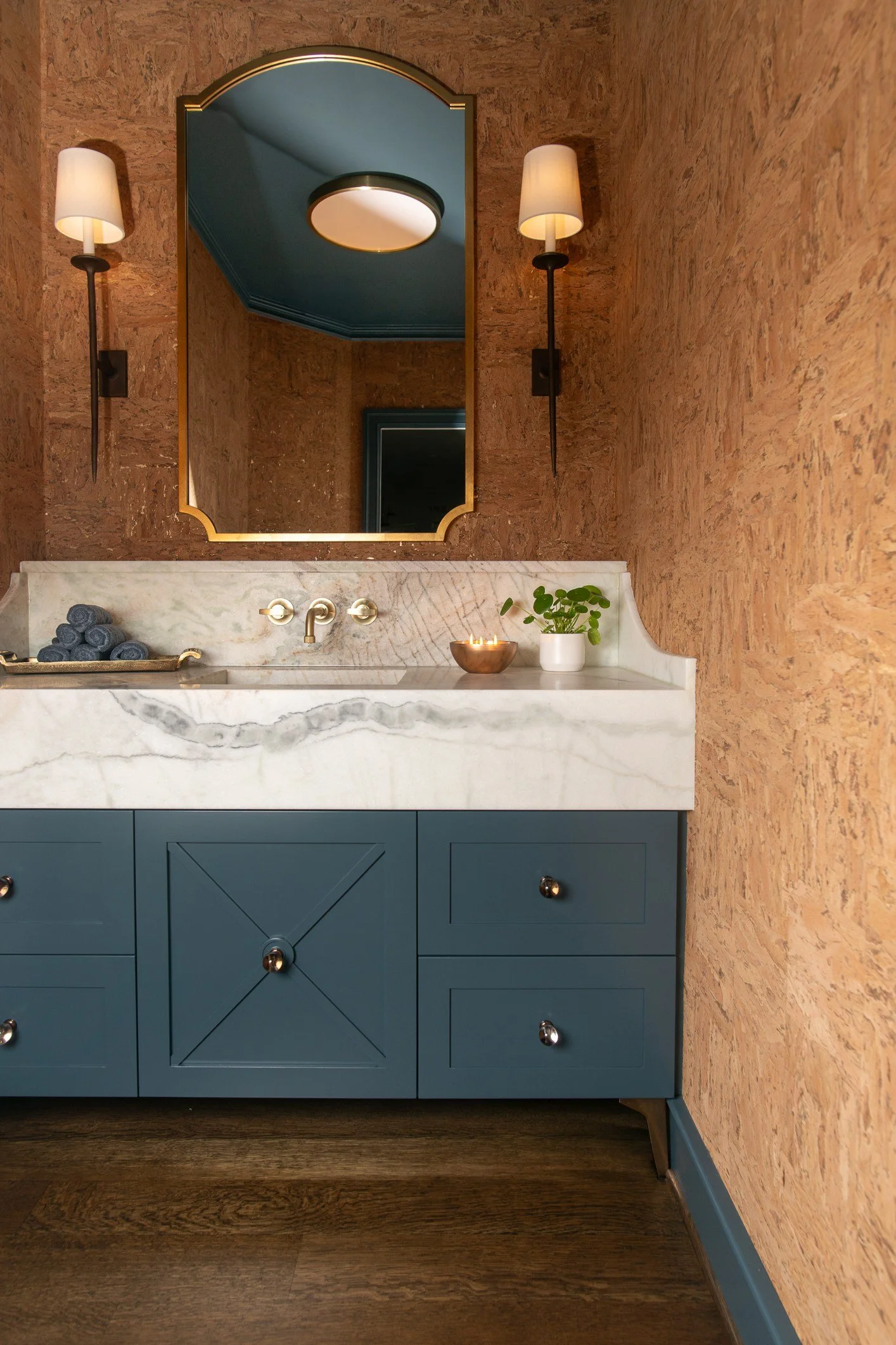 Bathroom vanity with blue cabinet, marble countertop, a large mirror, wall-mounted sconces, and decorative items including towels, a plant, and a bowl with candles.