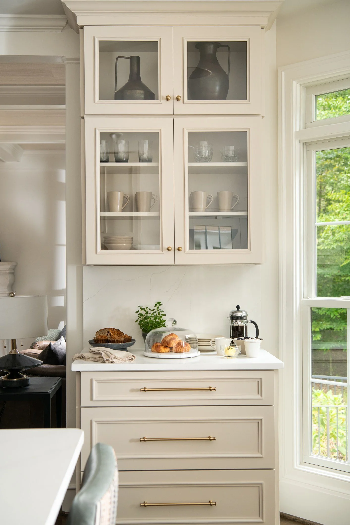 A kitchen cabinet with glass doors displaying white mugs and glassware, on a countertop with croissants, a French press, and cups.