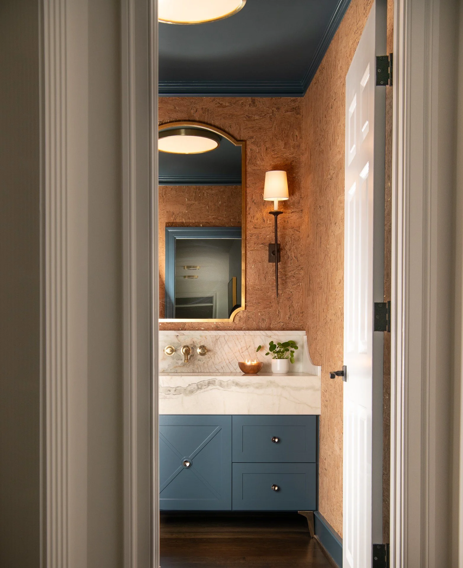 View of a small bathroom vanity with a blue cabinet, white marble countertop, a mirror, a wall-mounted lamp, and a small potted plant on the countertop. The walls are covered in a textured, reddish-brown material, and the ceiling is painted blue.
