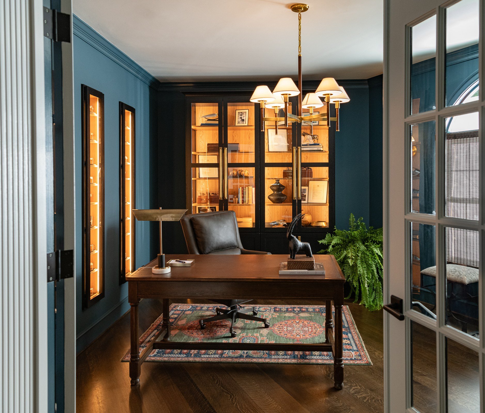 A well-decorated study or office with a wooden desk, leather chair, and built-in bookshelves. The walls are painted blue, and there's a chandelier hanging from the ceiling. The room has a rug on the wooden floor and a large green fern plant in the co