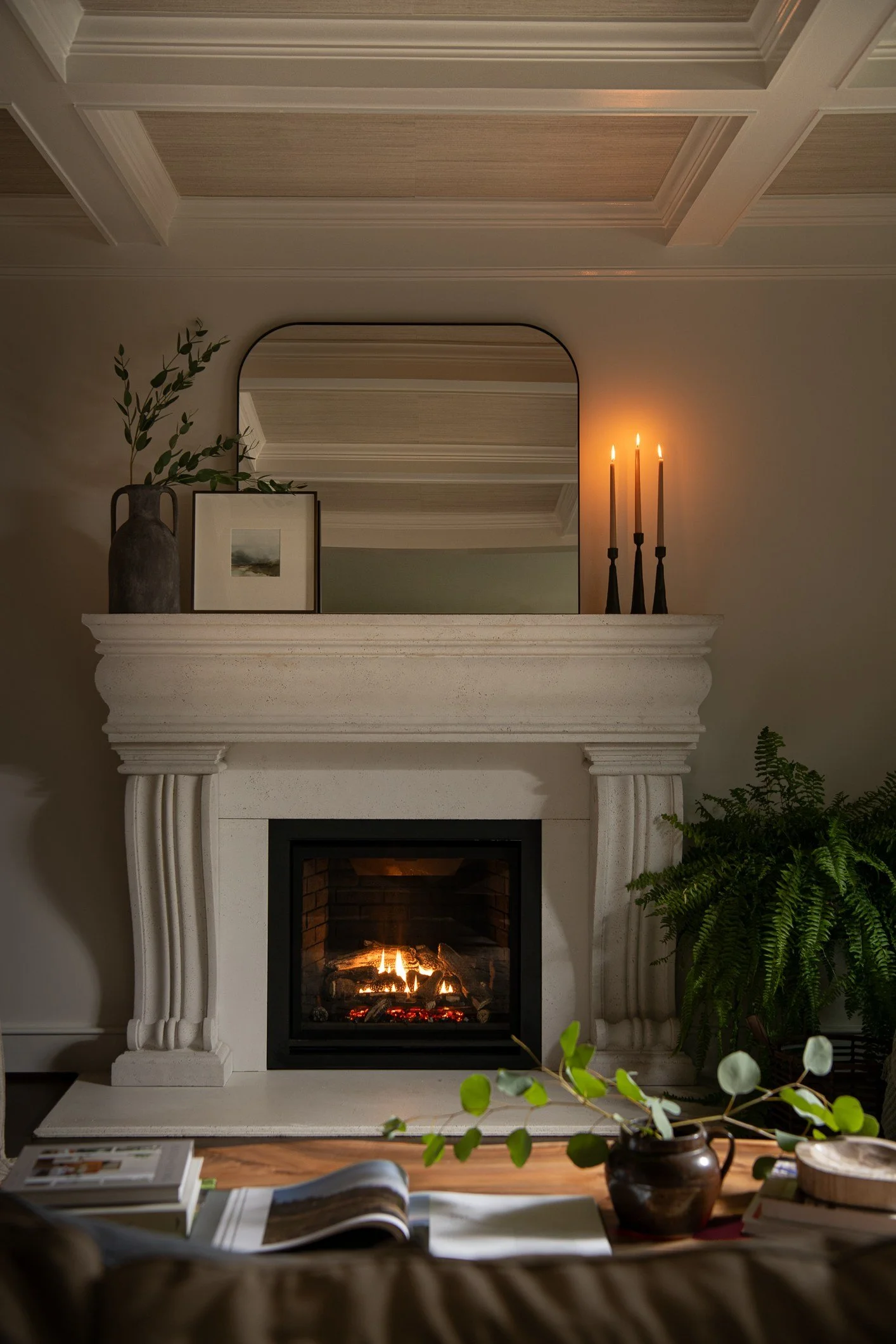 A cozy living room corner featuring a lit fireplace with a white mantel, decorated with a vase with greenery, a framed picture, and three lit candles. A mirror above reflects the ceiling, and a large potted fern is next to the fireplace. Part of a ta
