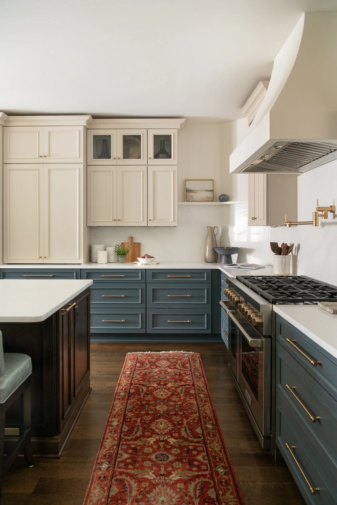 A kitchen with blue lower cabinets, white upper cabinets, a red patterned runner rug, and dark wood flooring.