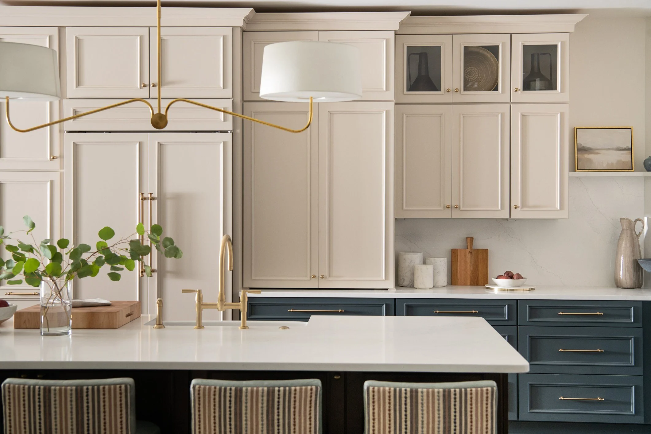 Modern kitchen with white upper cabinets, dark blue lower cabinets, a white countertop island, gold fixtures, and decorative items like vases, bowls, and framed art.