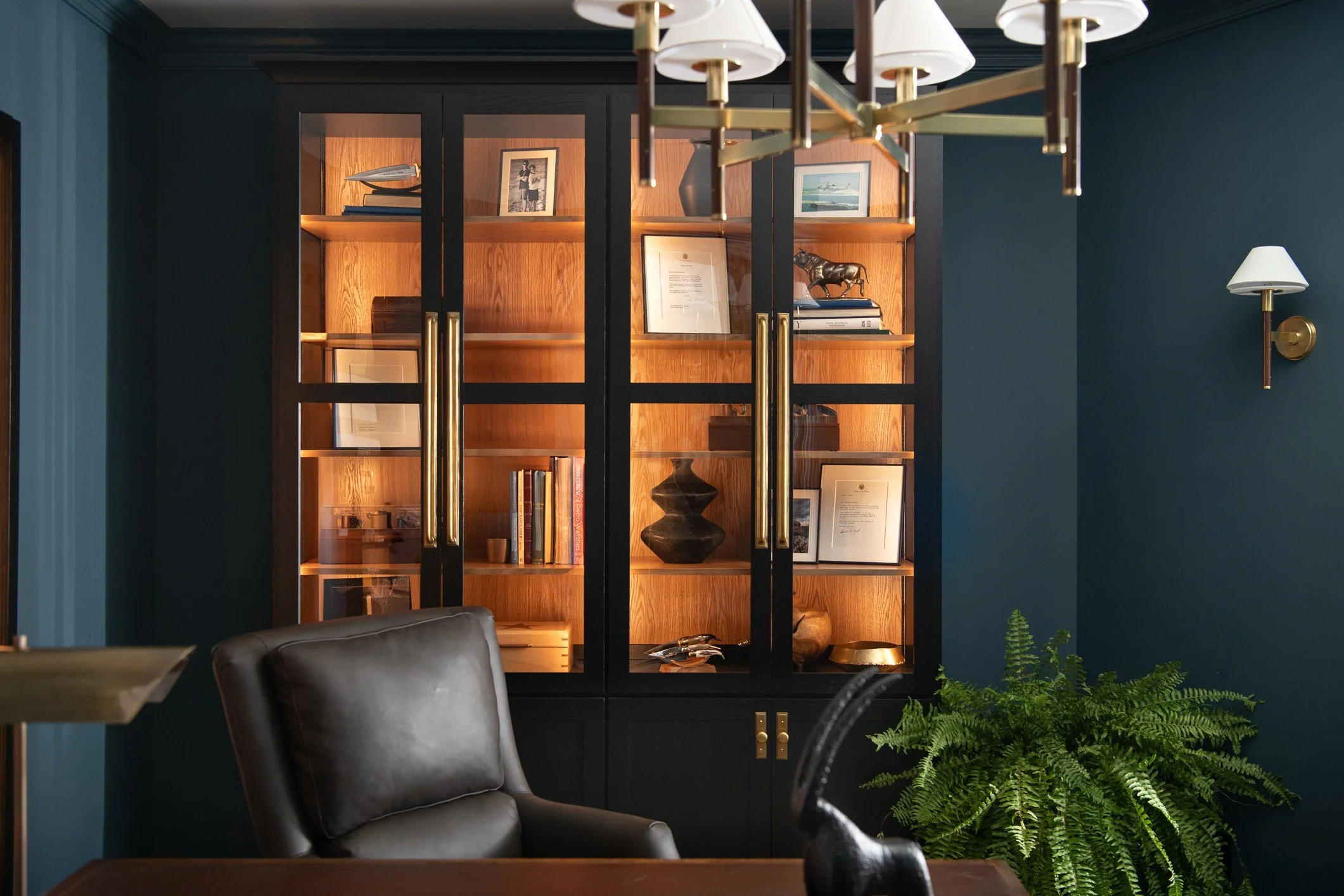 A room with a black and wood glass cabinet filled with books, framed certificates, and decorative items, a dark leather chair, a large green fern plant, and a modern chandelier and wall sconce lighting.