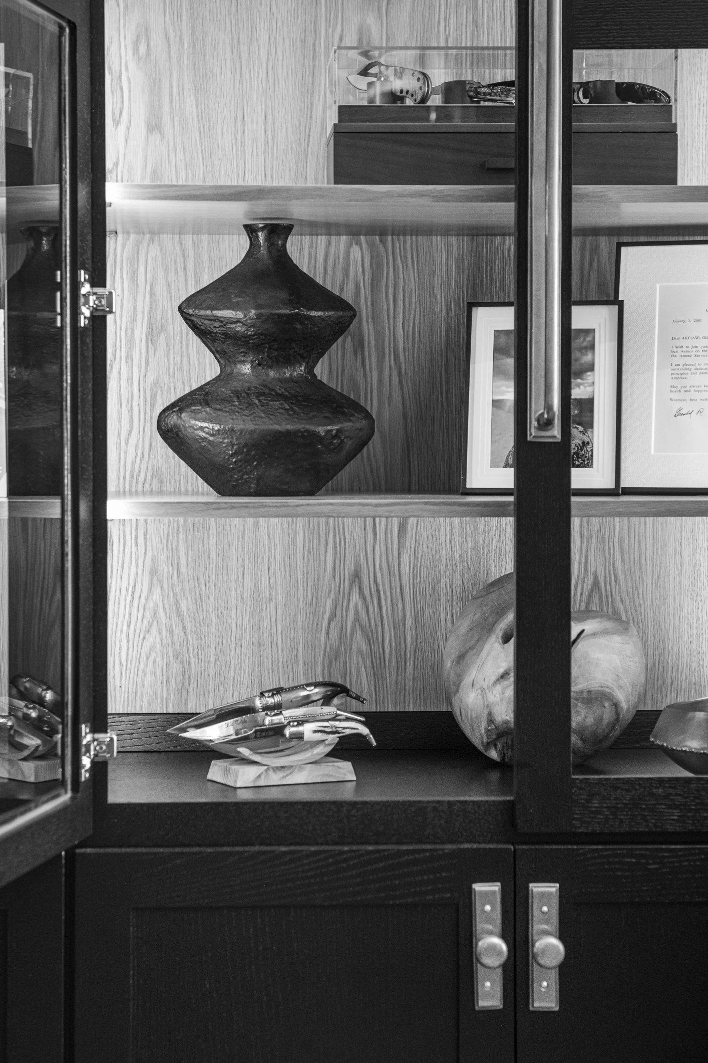 A black and white photo of a display cabinet with decorative items inside, including a dark ceramic vase, framed pictures, a wooden sculpture, and a metallic sculpture of hands.