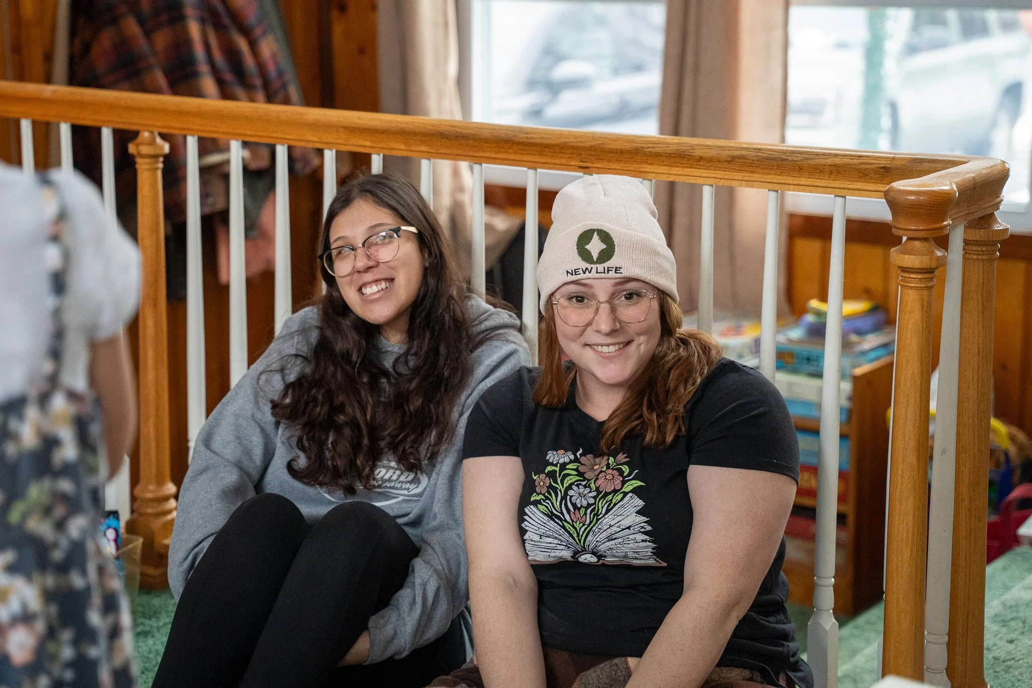 Two women sitting on the floor in front of a wooden railing, smiling. One woman is wearing glasses, a gray hoodie, and has long dark hair. The other woman has glasses, a beige beanie with a green logo and the words 'NEW LIFE', and a black T-shirt with a floral and open book design.