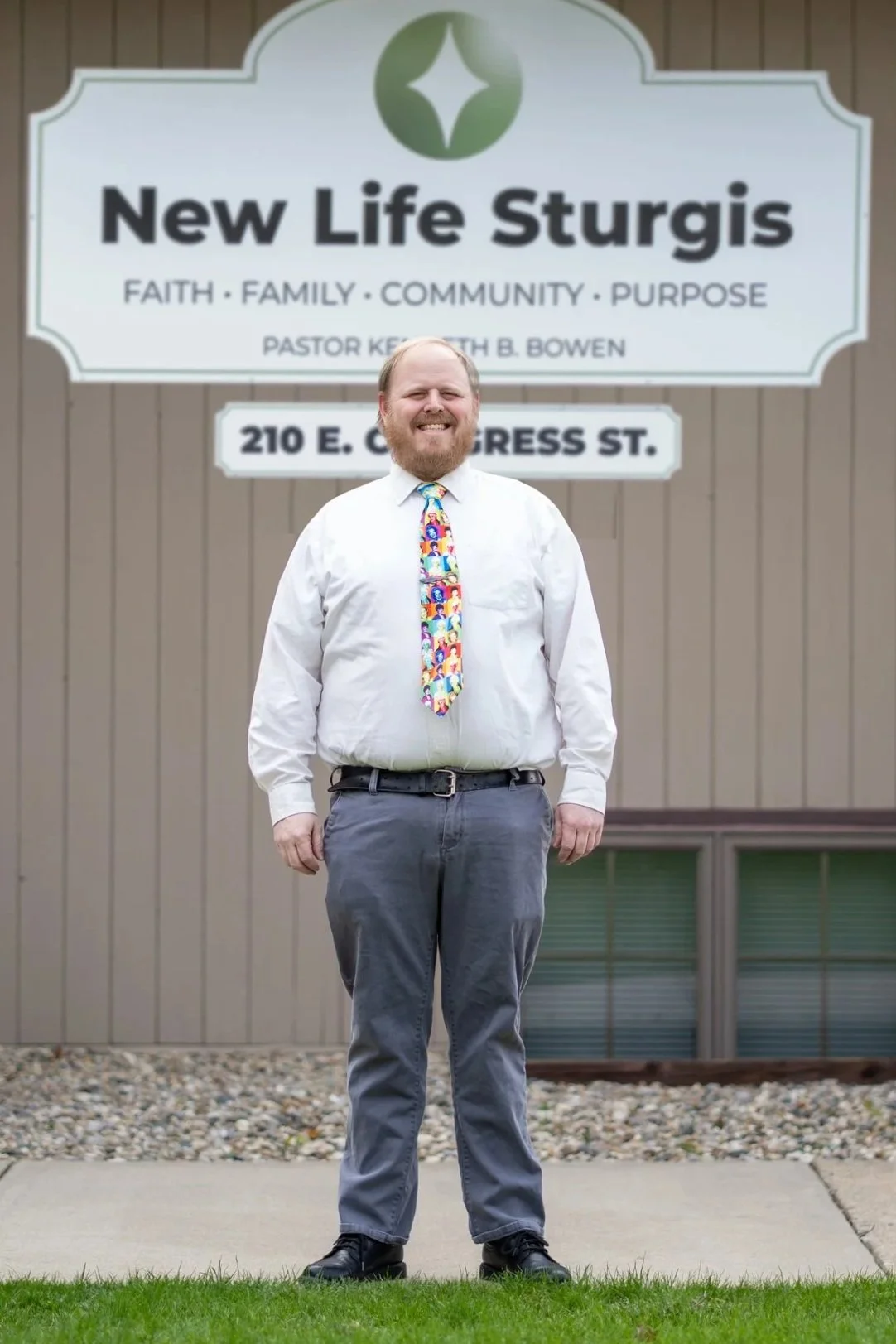 A person in a white shirt and colorful tie standing outside Potter's House Church of Sturgis with a sign displaying the church's name and address.
