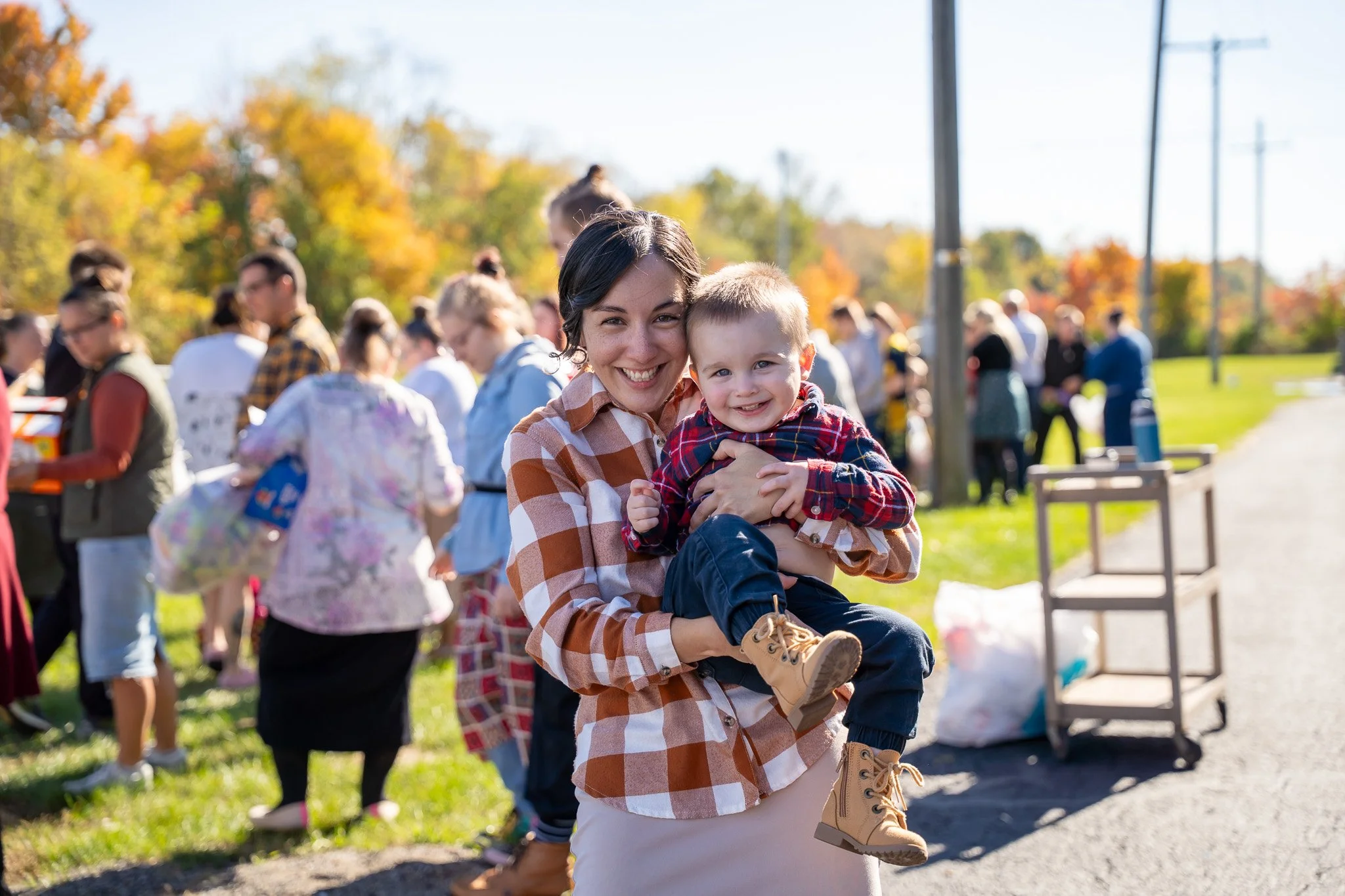 Smiling woman holding a young boy outdoors at a fall event with a crowd background and autumn trees.
