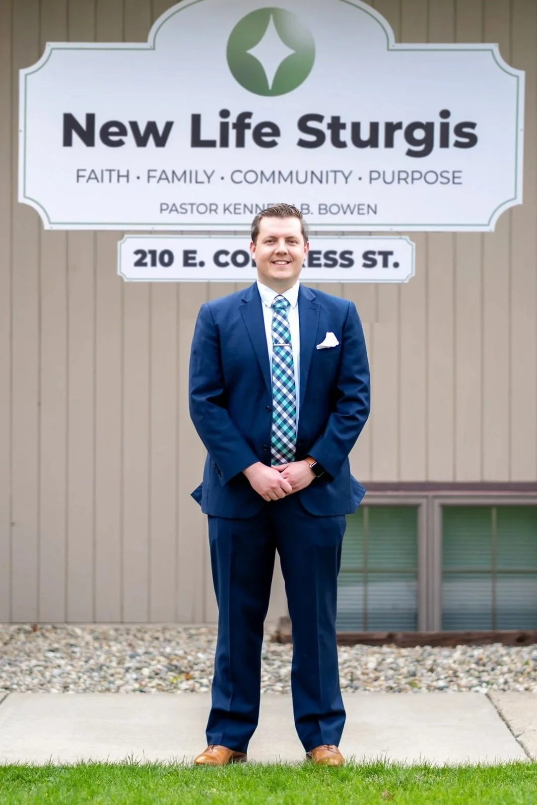 A man in a blue suit and checkered tie standing in front of Potter's House Church of Sturgis sign.
