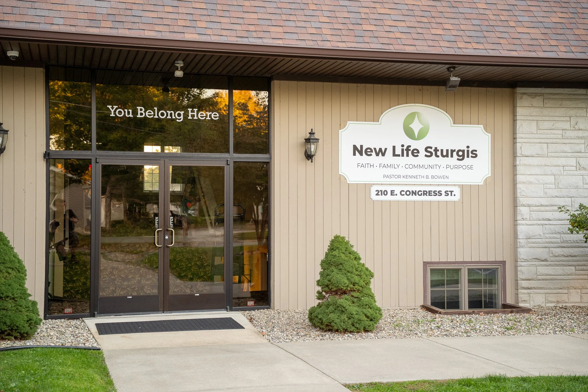 Exterior of a building with a beige wooden wall and a large white sign that reads 'New Life Sturgis' with the address 210 E. Congress St., and a glass door with the phrase 'You Belong Here' written on it. There are small bushes and gravel landscaping in front of the building.