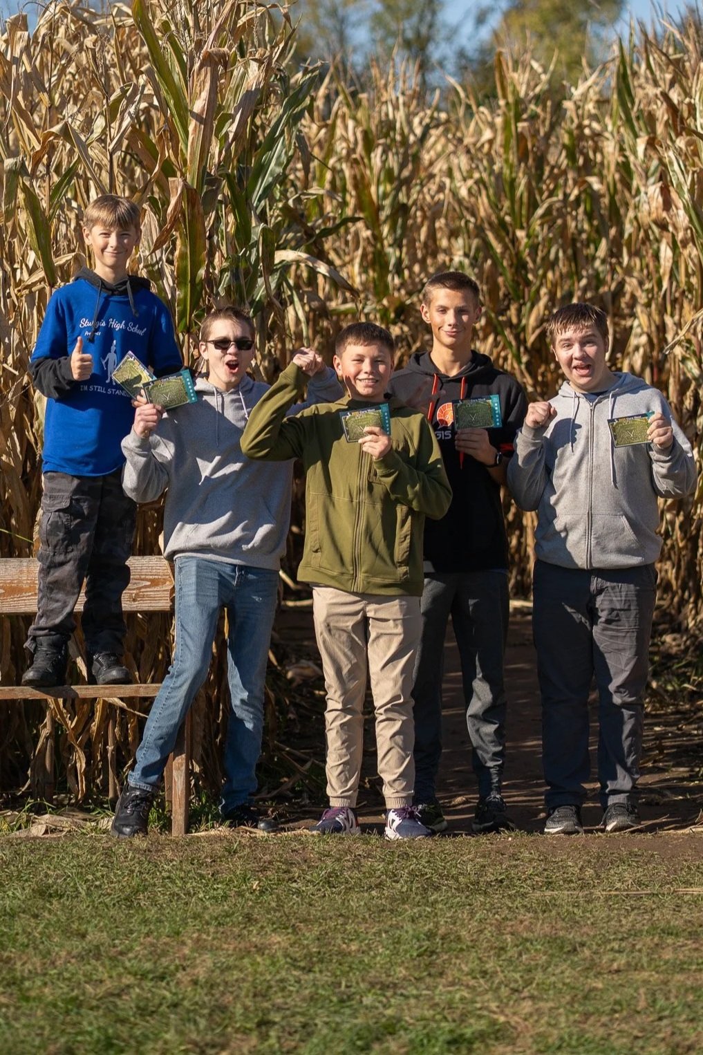 Five boys standing in front of a cornfield, holding yellow snack packages, and smiling. They are dressed in casual jackets and hoodies, with some giving thumbs-up or flexing their arms.