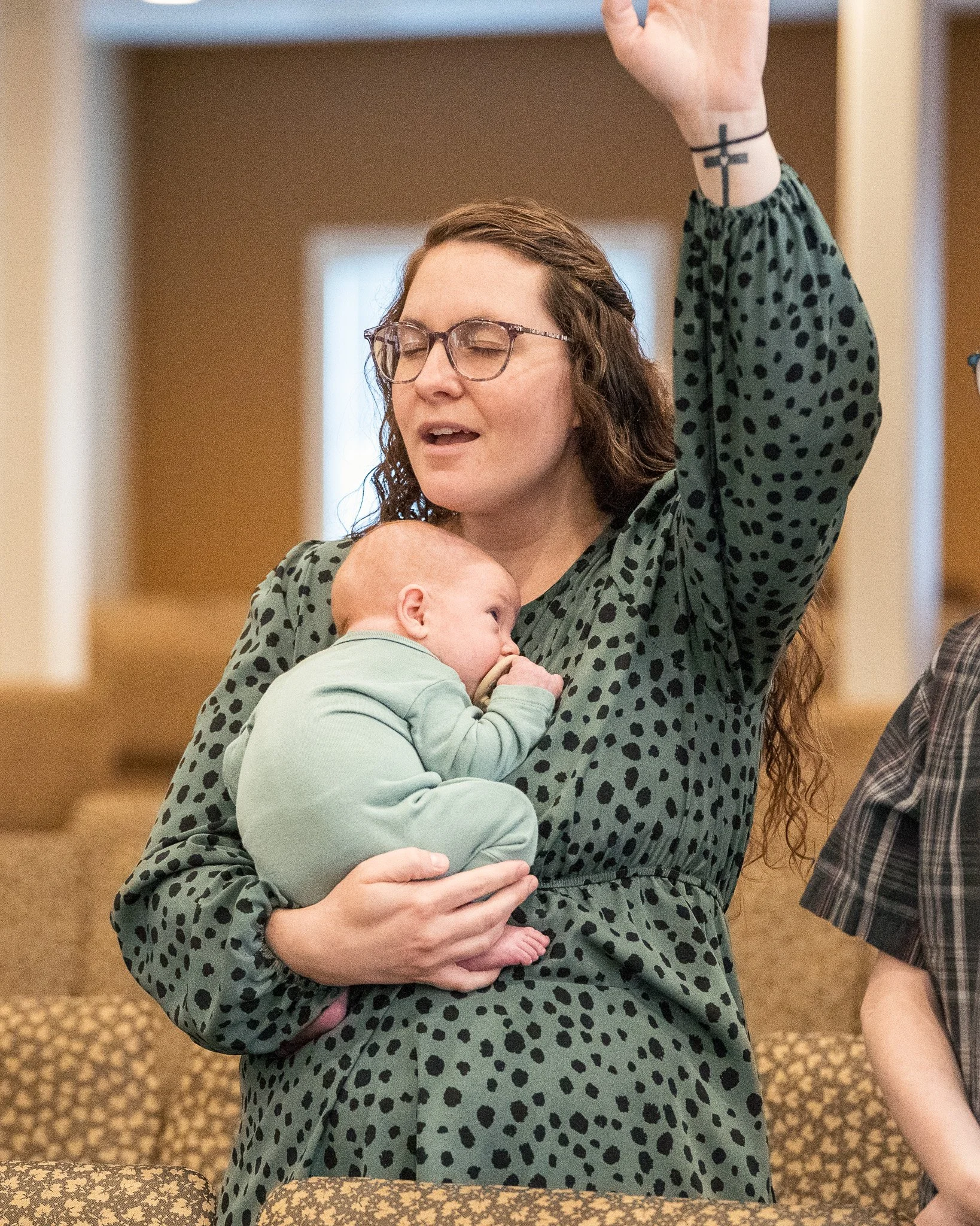 A woman with glasses and a cross tattoo on her wrist is holding a sleeping baby wearing a green onesie in her arms with her eyes closed and mouth slightly open, raising her left hand.