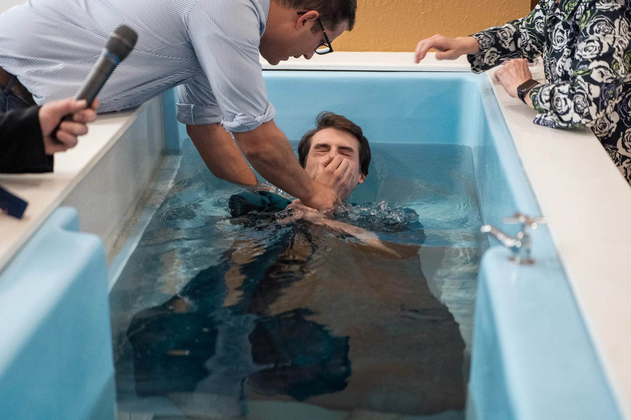 A man is being baptized in a baptismal pool by a man holding his nose, while a woman reaches towards him from the side.